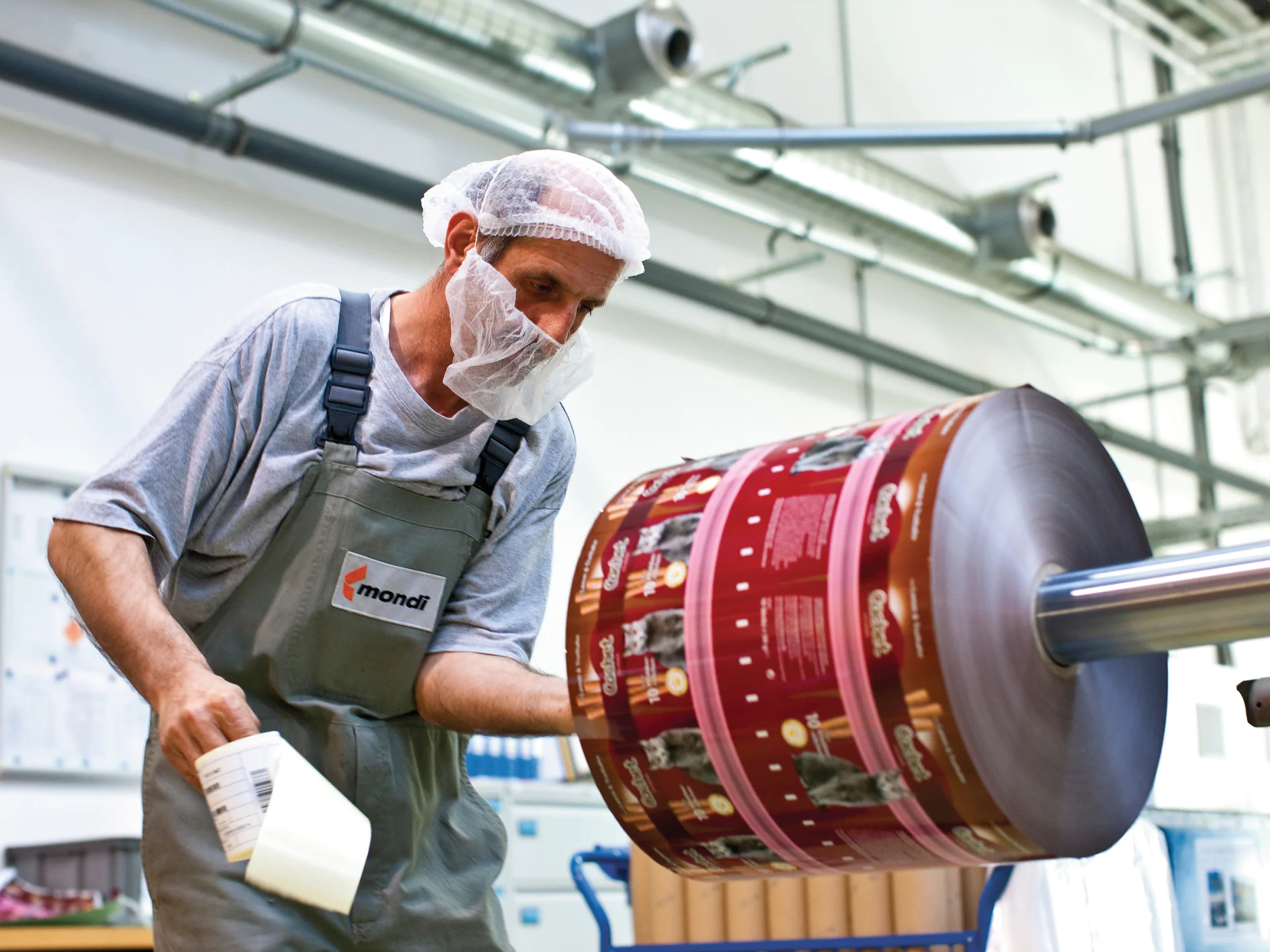 A worker wearing a hairnet and face mask inspects a large roll of printed packaging material in a factory setting. He holds a tool and is dressed in a gray shirt and green overalls. Machinery, tesa tape, and industrial elements are visible in the background. (This text has been generated by AI)