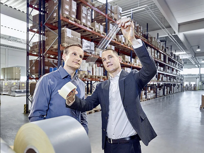Dos hombres examinan un rollo de material en un almacén; uno lo sostiene mientras el otro observa. El espacio está lleno de estanterías llenas de cajas y suministros. El entorno está bien iluminado, lo que sugiere un entorno industrial o de almacenamiento. (This text has been generated by AI)