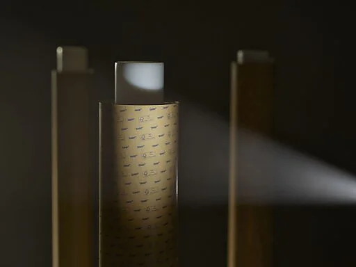 Tres rollos de cinta de embalaje marrón de Tesa están dispuestos verticalmente en una zona con poca luz, con el rollo central ligeramente iluminado por un haz de luz. El primer plano está enfocado mientras que el fondo permanece oscuro y borroso. (This text has been generated by AI)