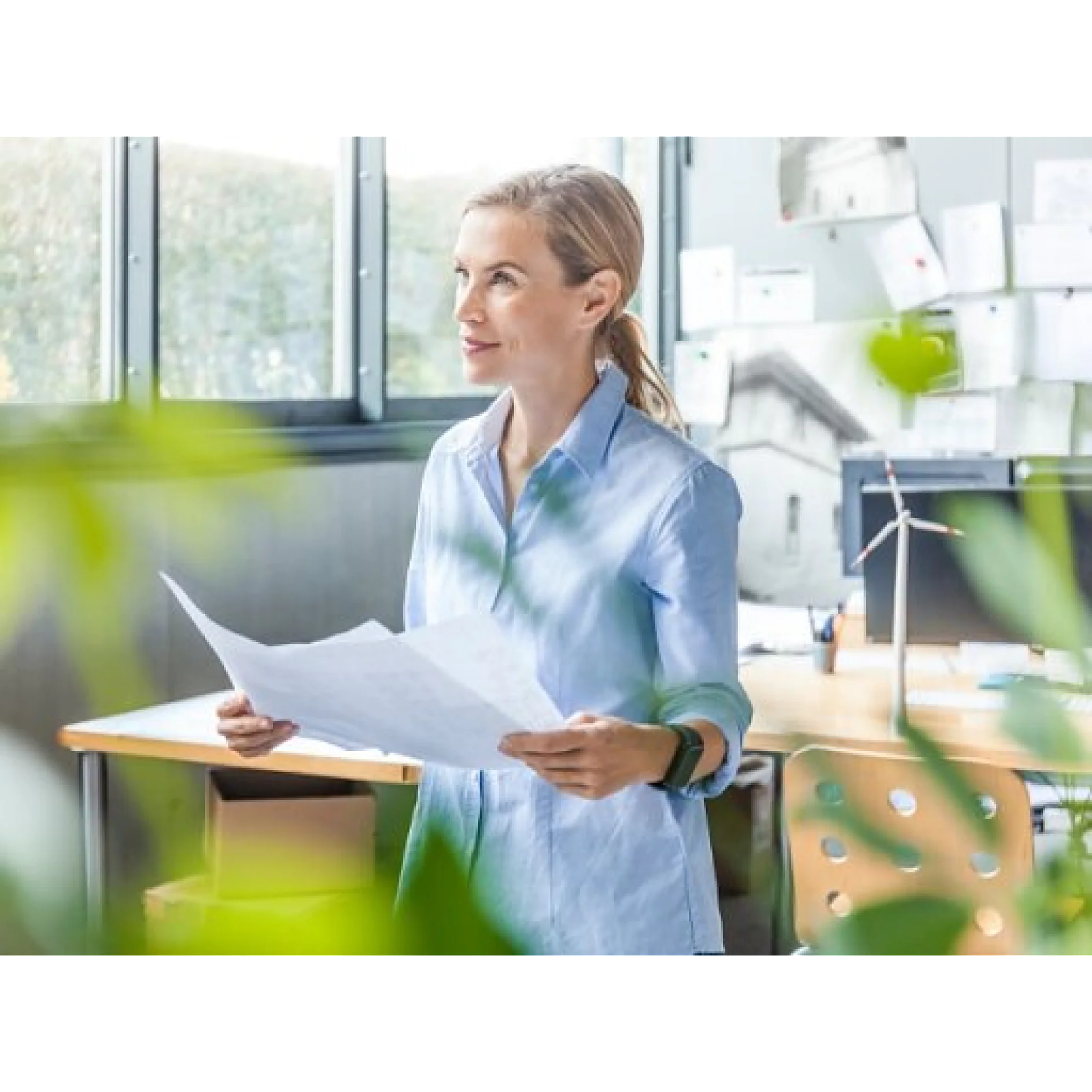 En una oficina, una persona con una camisa azul claro sostiene papeles. La habitación cuenta con grandes ventanales, un escritorio de madera y varios papeles y cuadros en las paredes. En primer plano se ve parcialmente una planta. (This text has been generated by AI)