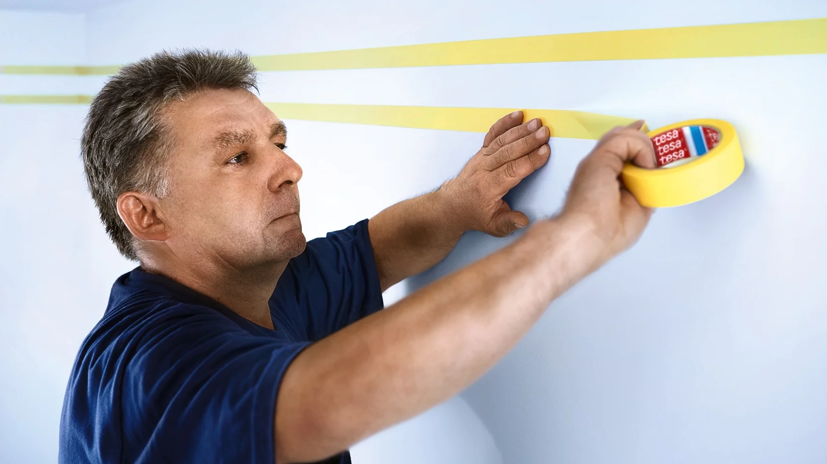 tesa_professional_masking_4334_step_1of3_ap-Print Un hombre con camisa azul aplica cinta tesa amarilla en líneas rectas sobre una pared azul claro, preparando la superficie para pintar. (This text has been generated by AI)