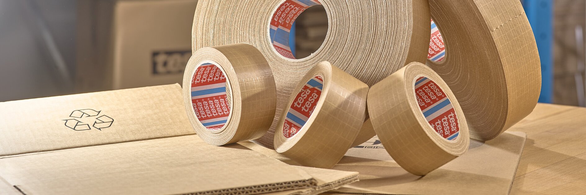 Varios rollos de cinta tesa de embalaje marrón de diferentes tamaños se exhiben sobre una mesa. Al fondo, hay cajas y paquetes de cartón apilados en estanterías azules. En primer plano, hay una caja de cartón con un símbolo de reciclaje.