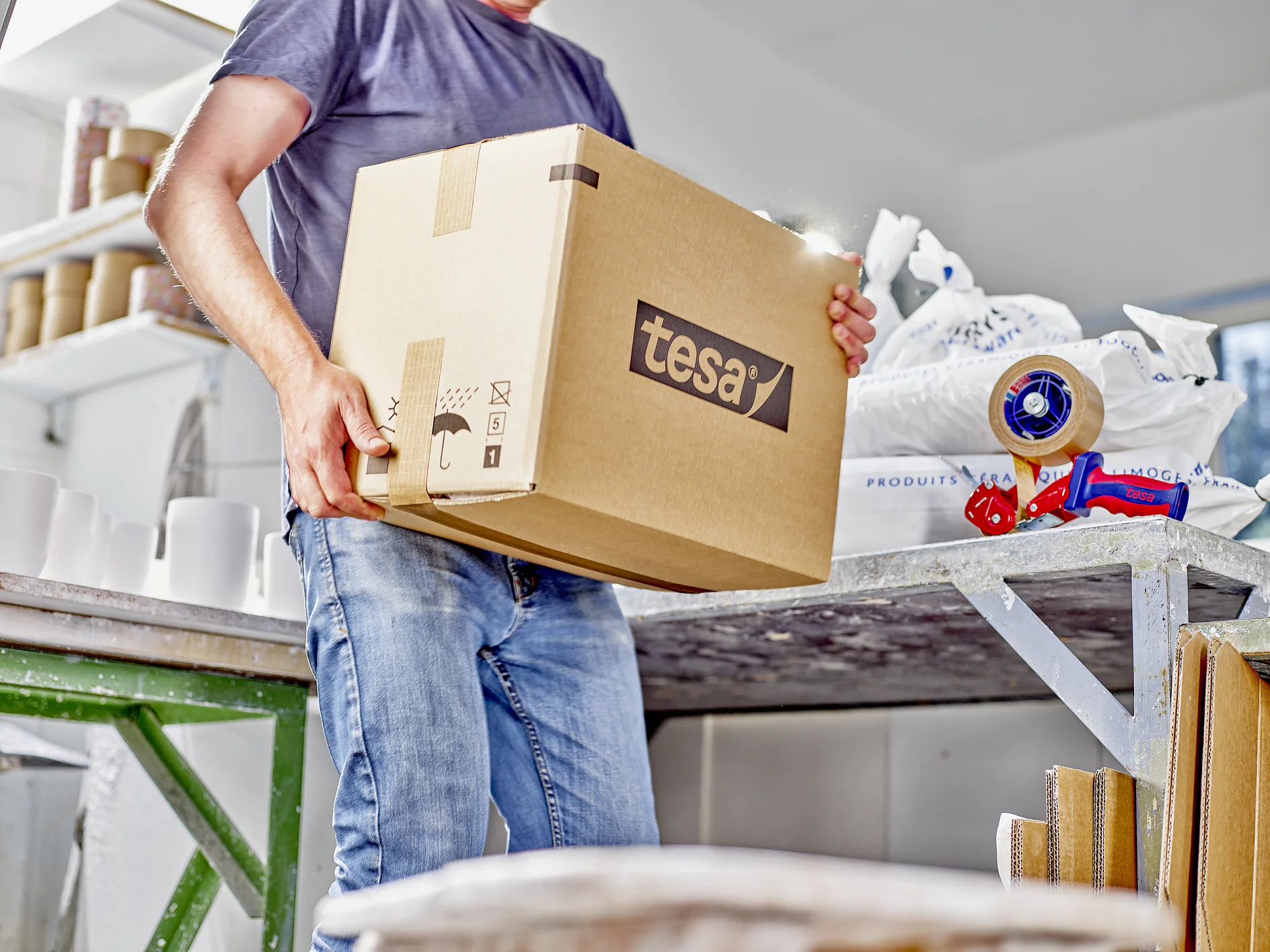 Una persona vestida con jeans y una remera gris lleva una caja de cartón grande etiquetada como tesa en un espacio de trabajo. Cerca, hay una mesa con materiales de embalaje, incluido cinta tesa y un dispensador. (This text has been generated by AI)