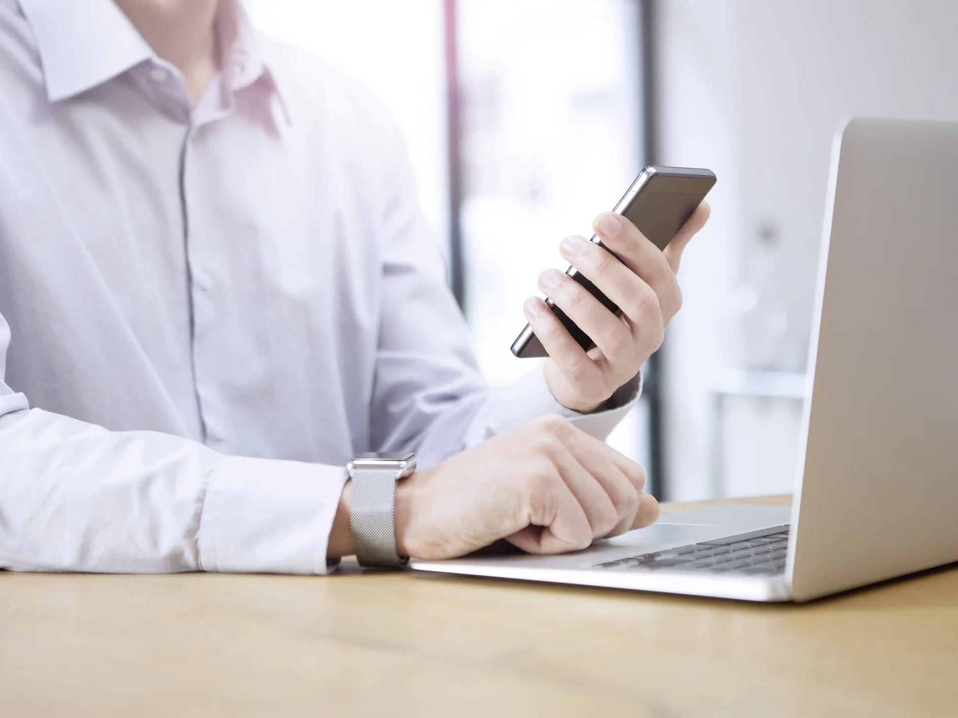 Una persona con una camisa blanca está sentada a una mesa usando un teléfono inteligente y una laptop. La persona sostiene el teléfono con la mano izquierda mientras la mano derecha descansa sobre la laptop cubierta con cinta tesa. El fondo está desenfocado con luz natural entrando. (This text has been generated by AI)