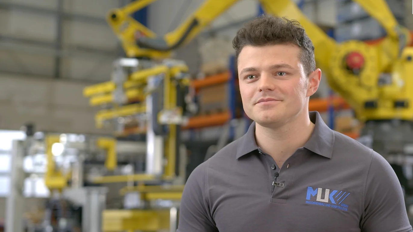 A man with short dark hair is wearing a gray polo shirt with a logo. He stands in an industrial setting with yellow machinery and blurred equipment in the background, surrounded by rolls of tesa tape. The atmosphere suggests a factory or manufacturing environment. (This text has been generated by AI)
