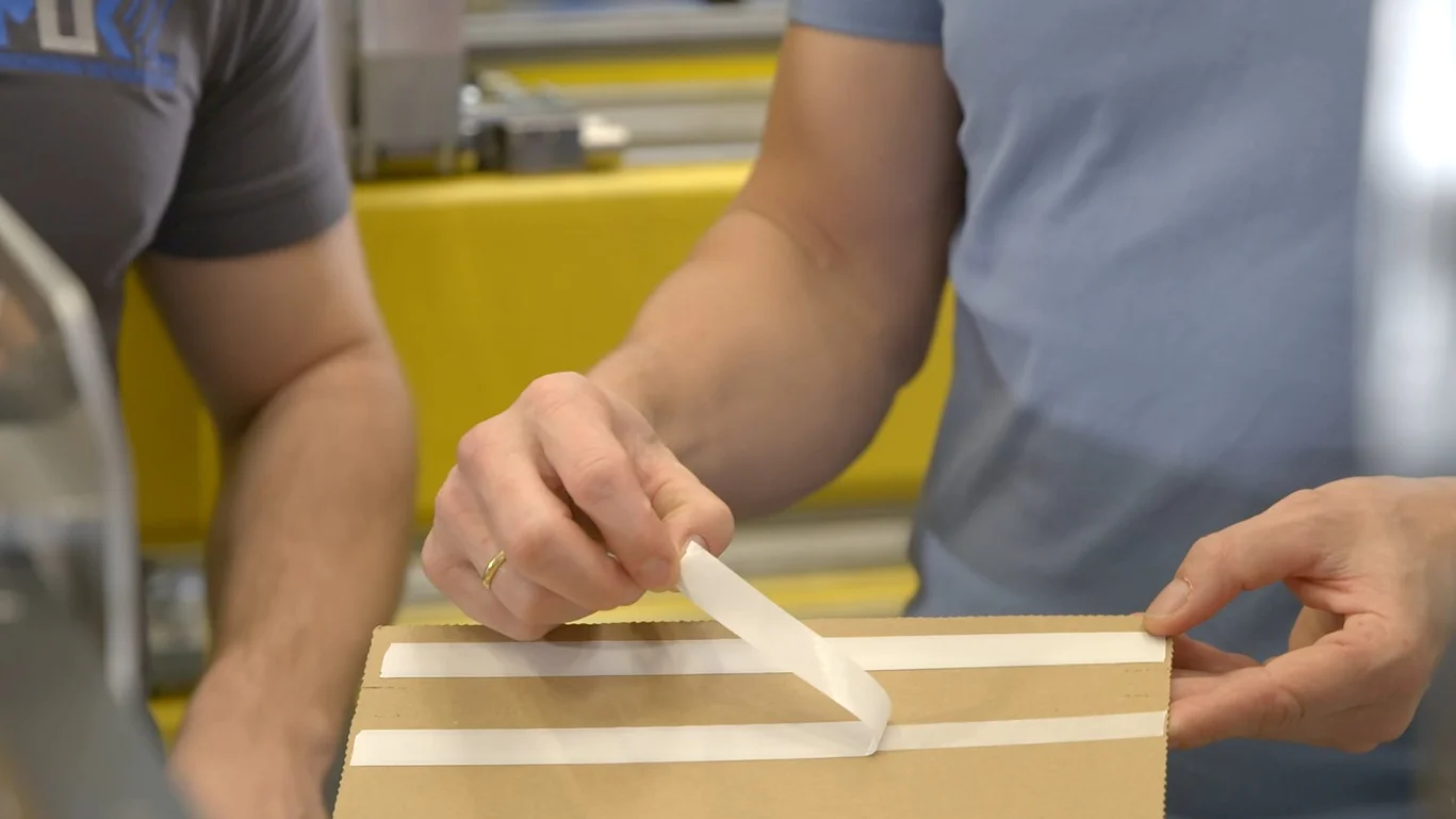 Two individuals are assembling a cardboard box, using white tesa tape to seal it. One person is peeling the tesa tape, while the other observes. They are in an indoor setting with a yellow background. (This text has been generated by AI)