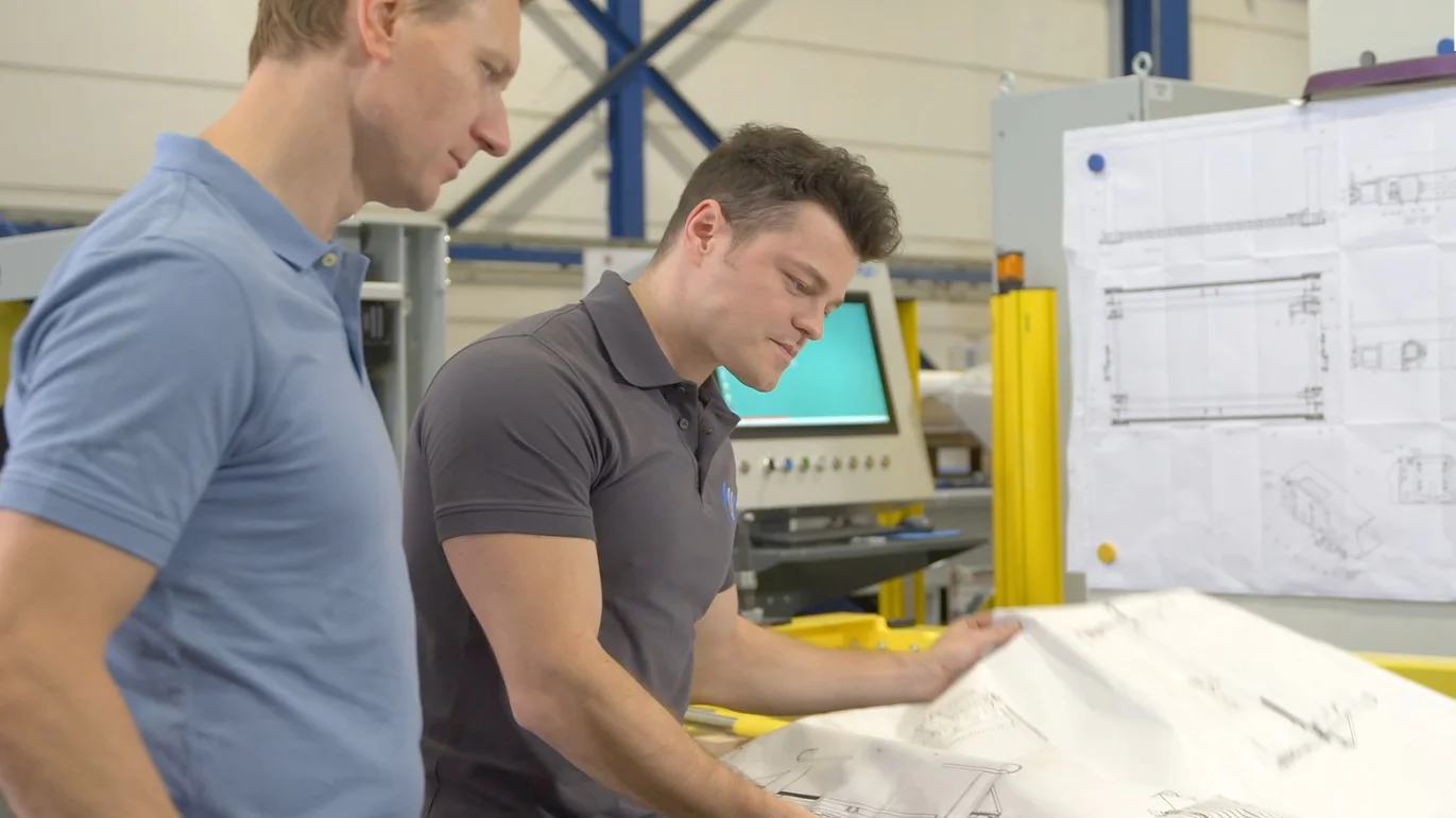 Two men in polo shirts examine engineering blueprints in an industrial setting. One holds the paper while the other observes. In the background, there are additional blueprints pinned to a board using tesa tape, with industrial equipment visible. (This text has been generated by AI)