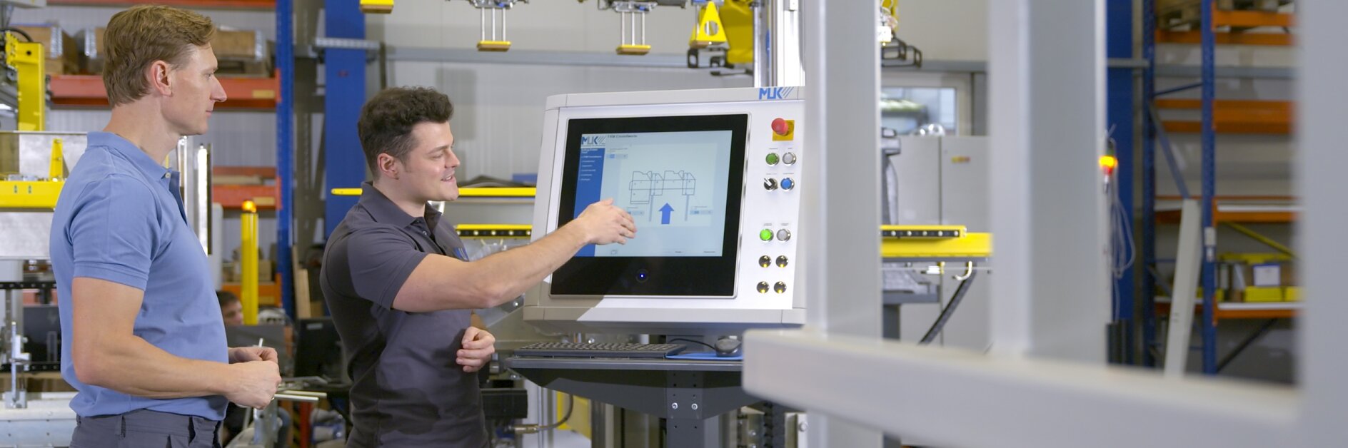 Two men stand by a control panel in an industrial setting. One man points at a diagram on a large monitor, while the other observes. Machinery with yellow components is visible in the background, indicating areas where tesa tape might be used. Both are dressed in casual work attire. (This text has been generated by AI)