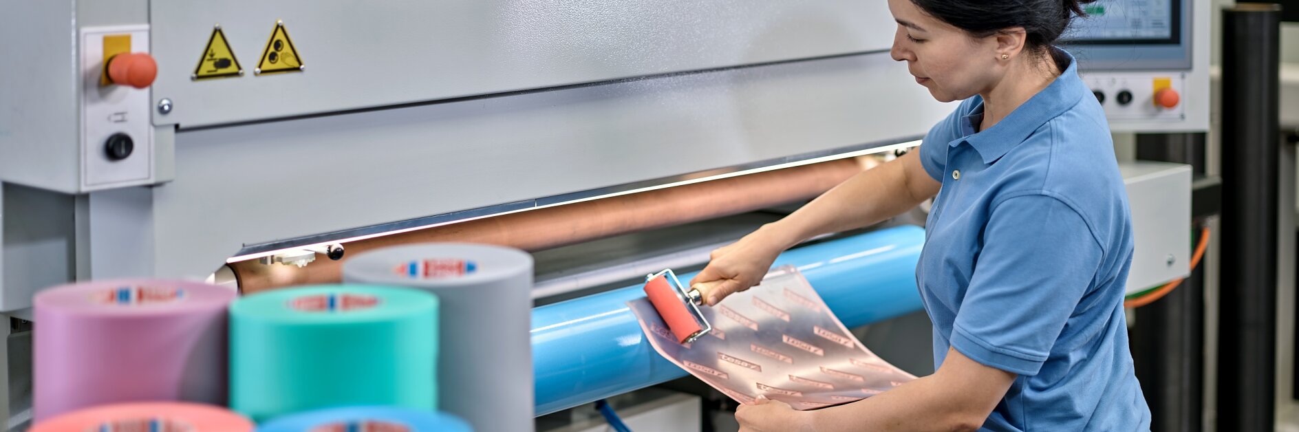 A woman in a blue shirt operates a large industrial printing machine. She is placing a patterned sheet onto a rotating drum. Rolls of variously colored tesa tape are visible in the foreground. A monitor displays information above her.