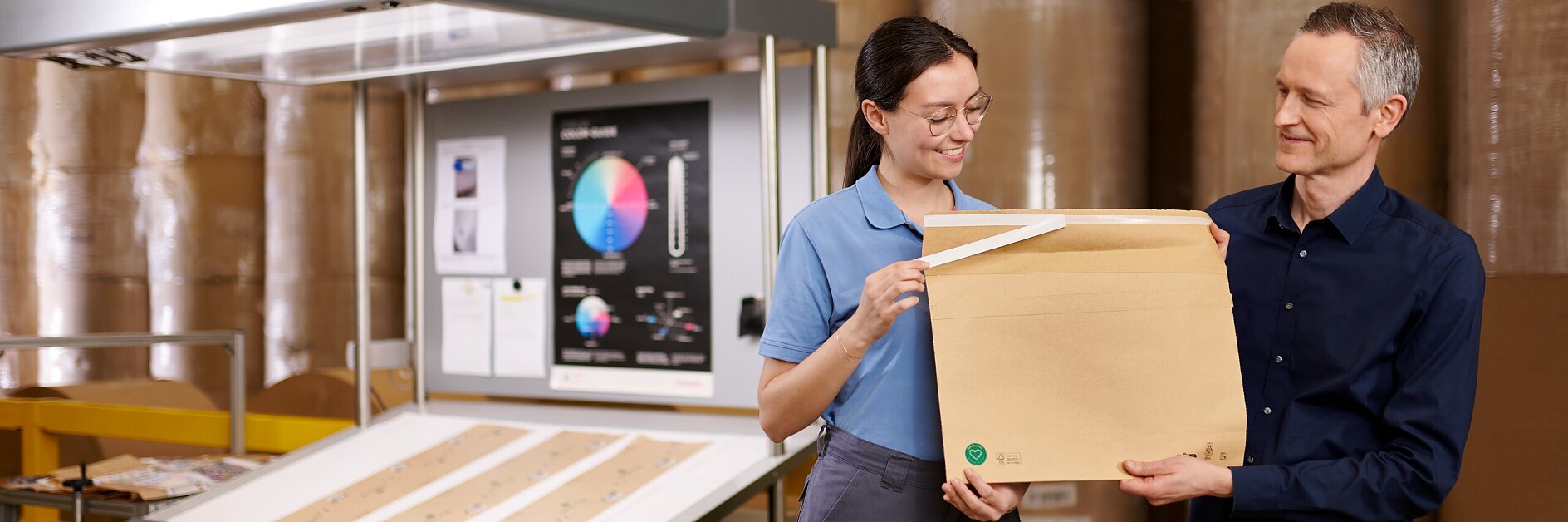 two workers in a warehouse holding a cardboard box near large rolls of paper and a printing machine labeled tesa
