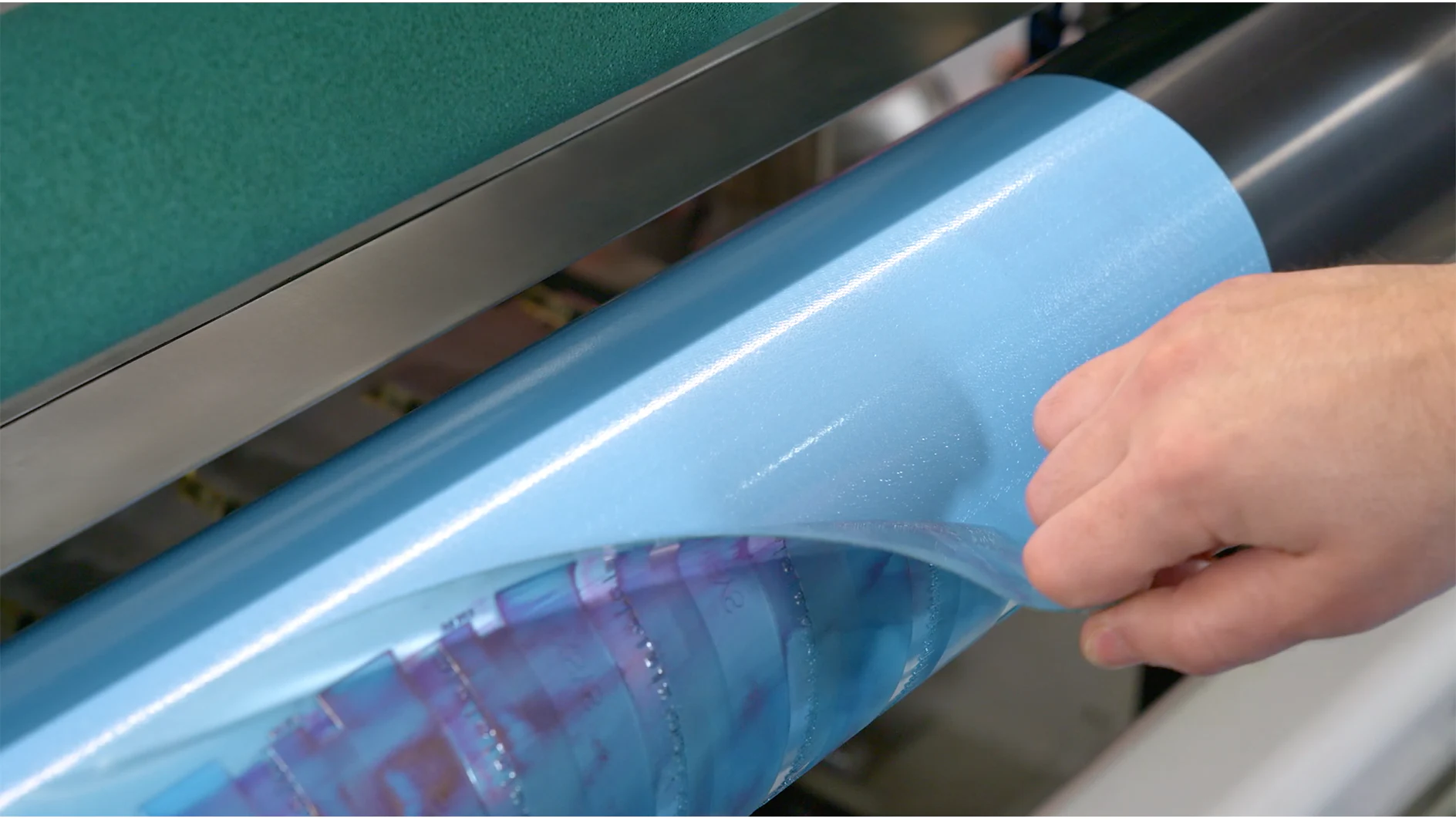 A hand peels a blue, glossy film from a cylindrical surface. The background includes blurred industrial machinery, and the photo focuses on the action of removing the tesa tape. (This text has been generated by AI)
