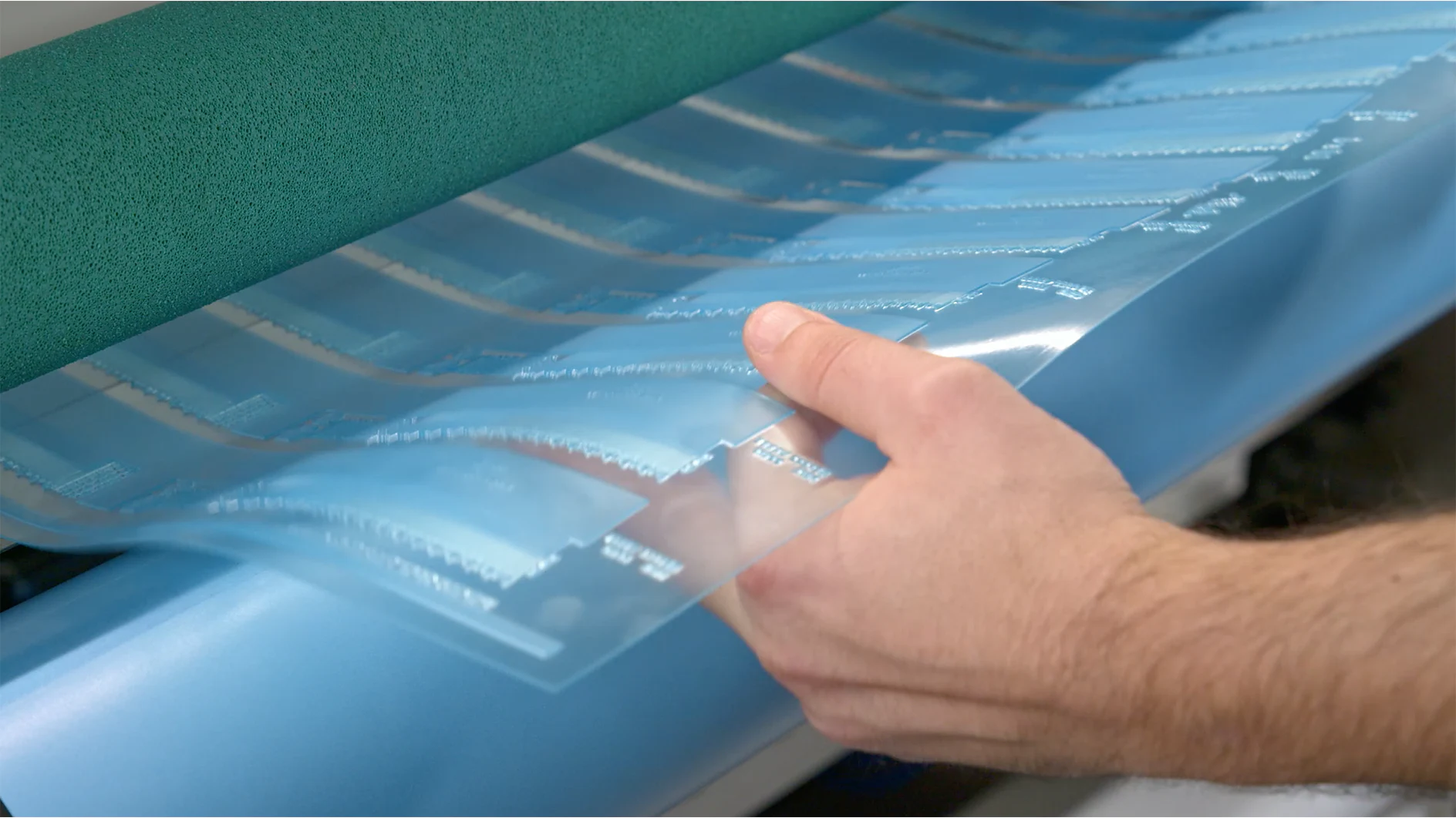 A person holds a blue sheet of photopolymer film with multiple horizontal tesa tape strips and text. The sheet is being fed into a printing or processing machine with visible rollers. The background is blurred. (This text has been generated by AI)