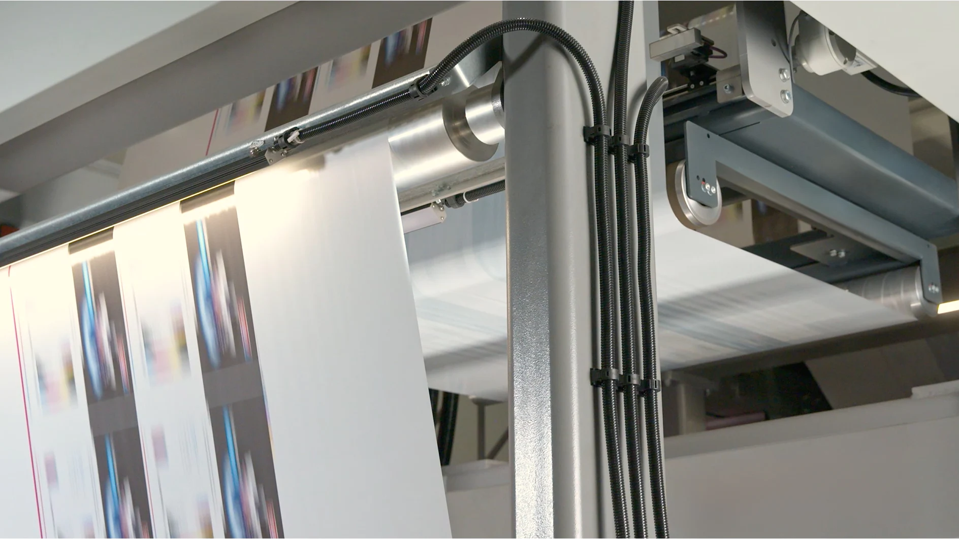 A printing machine processes sheets of paper, printing color images. The machines metallic components are visible, with wires running alongside it. A section of the printed paper is shown in motion, secured with tesa tape. (This text has been generated by AI)