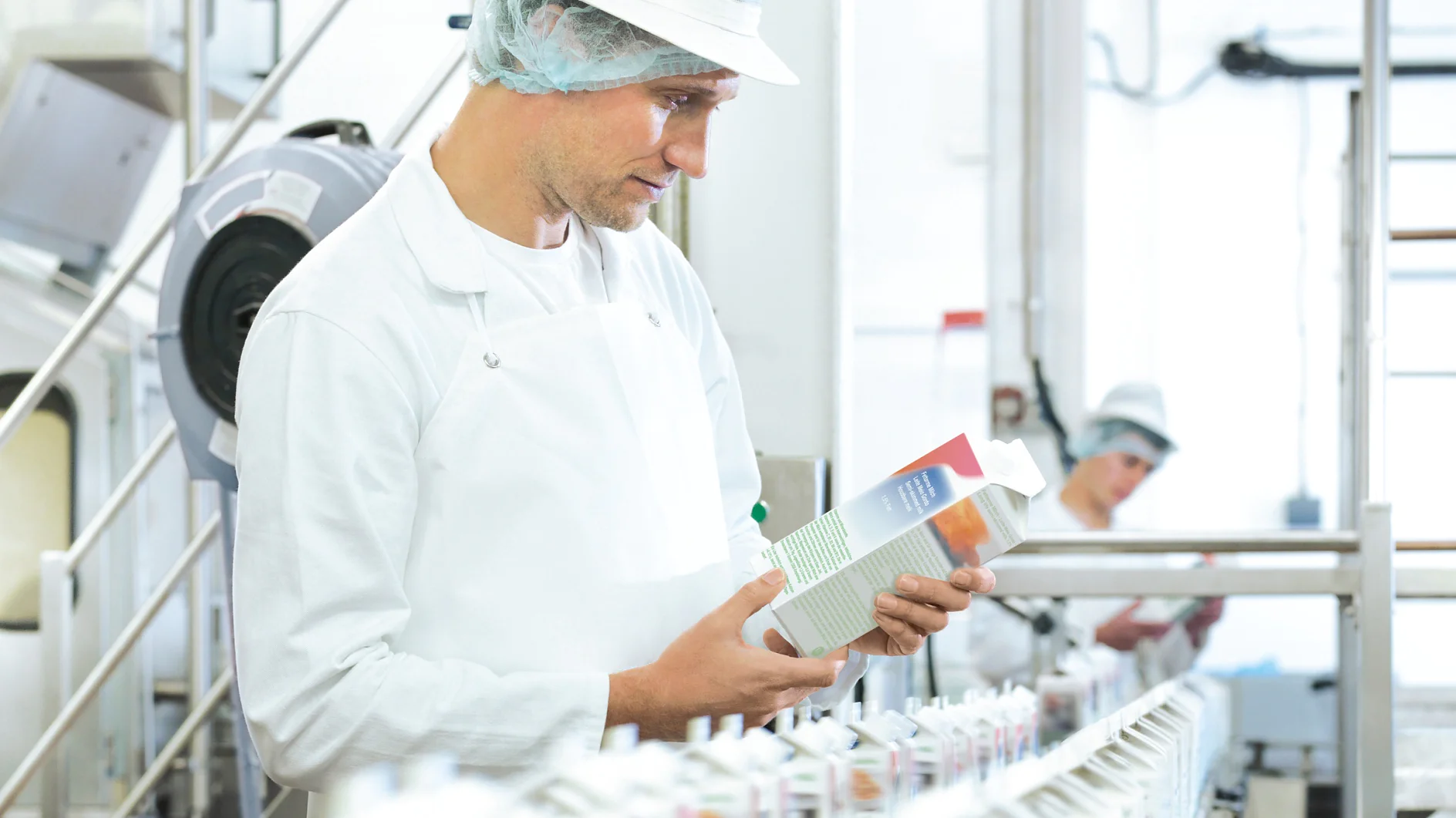 A factory worker in a white apron, hairnet, and cap inspects a carton on an assembly line filled with similar cartons sealed using tesa tape. Another worker in the background is also wearing similar attire in a well-lit, industrial setting. (This text has been generated by AI)