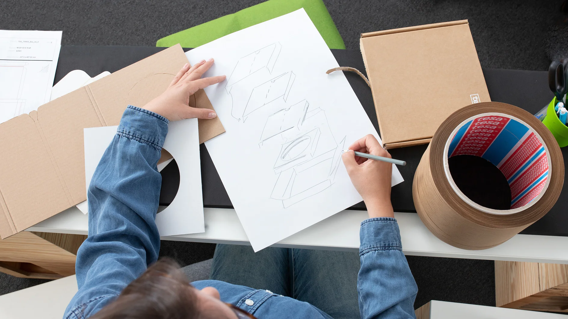 A person in a denim shirt is sketching a box design on white paper. A circular cardboard container, open cardboard box, and tesa tape are on the table. The background includes a black and white table surface with green material. (This text has been generated by AI)