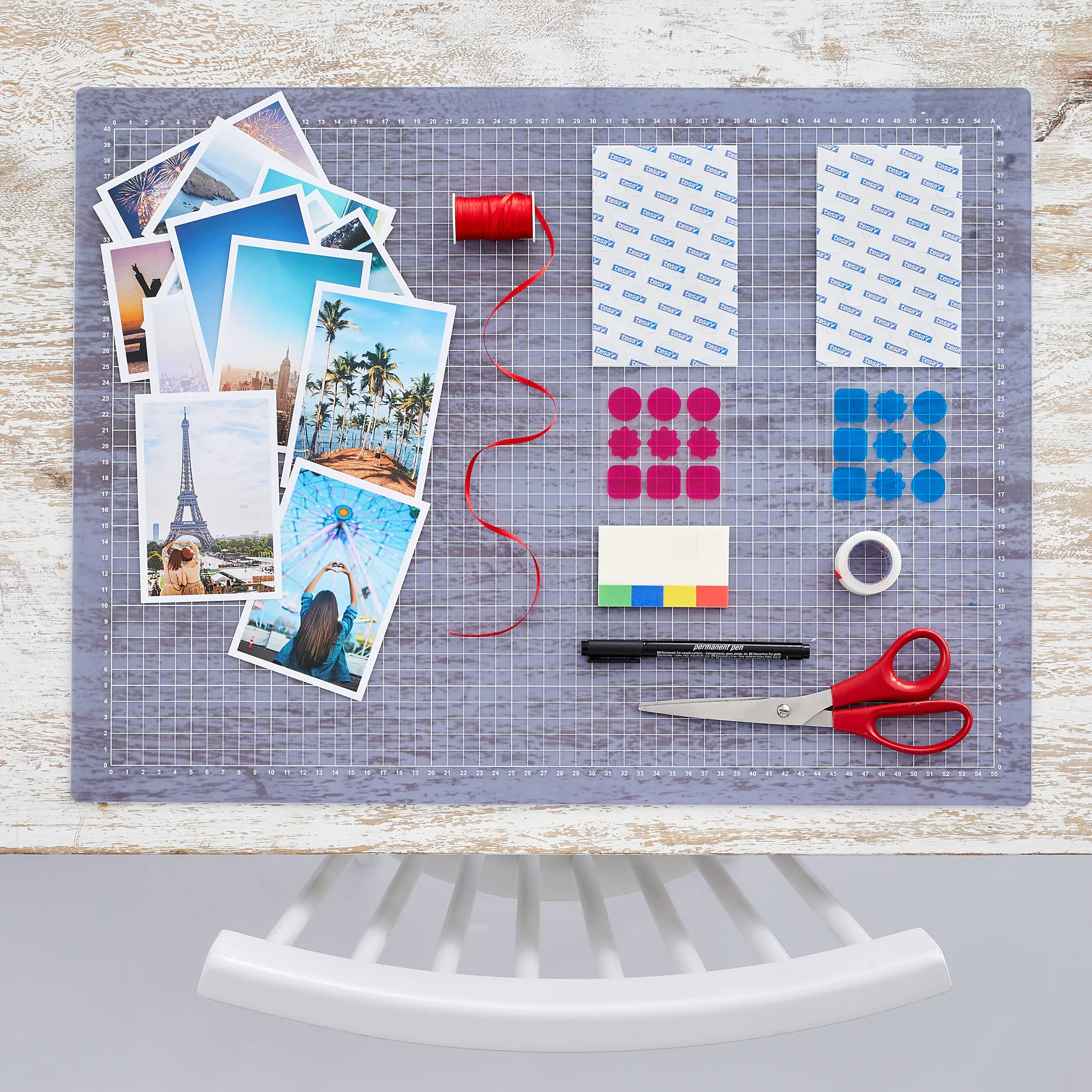 A flat lay of crafting materials on a tabletop includes photos, red thread, scissors, adhesive squares, colored stickers, a black pen, a tesa tape roll, and a white chair is partially visible at the bottom. (This text has been generated by AI)
