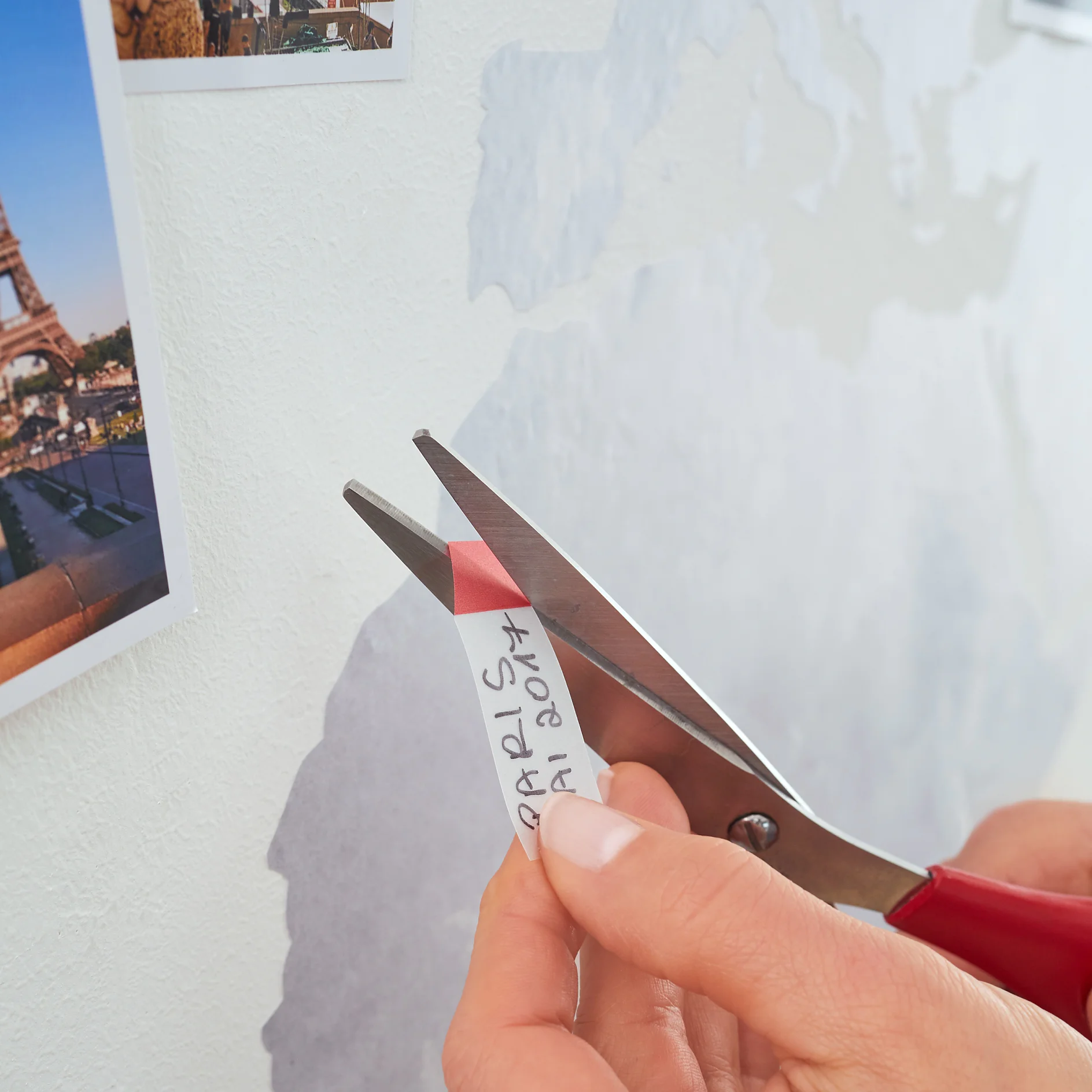 A person is using a pair of scissors to cut a strip of paper labeled PARIS 01.2017. The strip is being held against a wall with tesa tape. A photograph of the Eiffel Tower is partially visible in the background. (This text has been generated by AI)