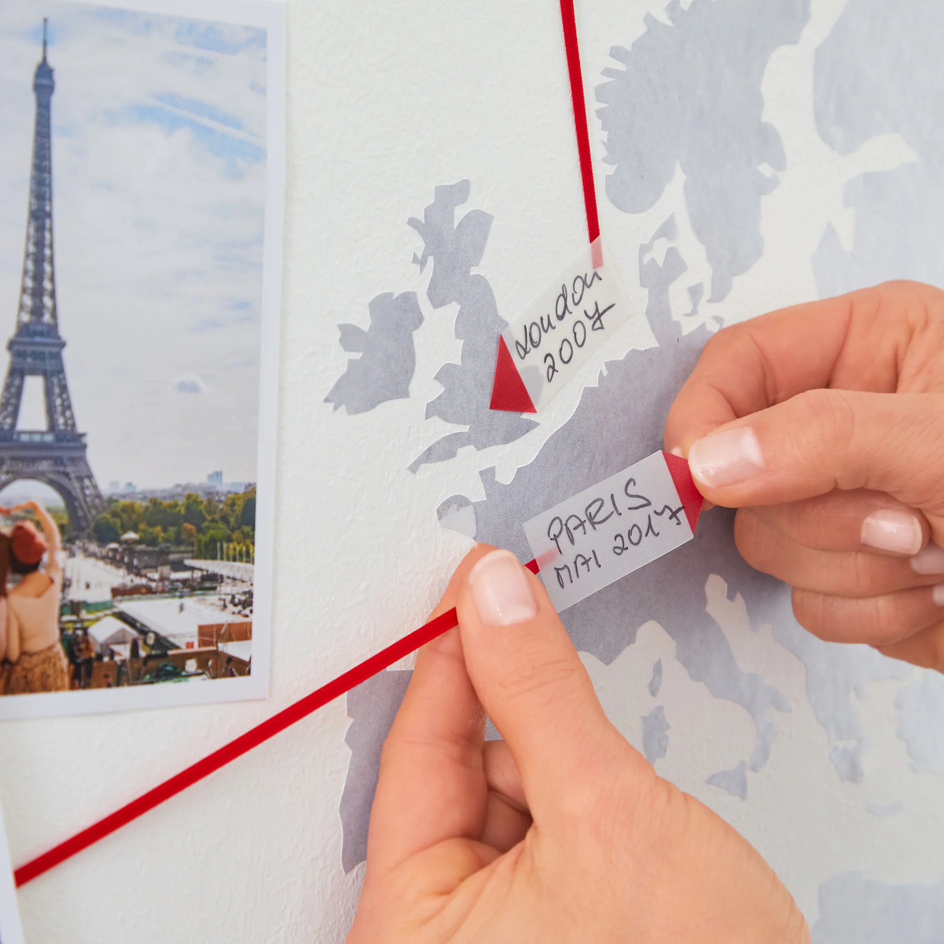 Close-up of hands pinning a paper note reading PARIS MAY 2017 on a map with red string connected to another note labeled Noulan 2004. A photo of the Eiffel Tower is on the left side of the map, secured using tesa tape. (This text has been generated by AI)