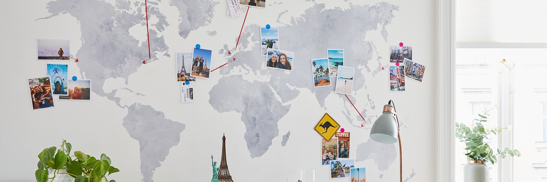 A world map on a wall is decorated with various travel photos connected by strings using tesa tape. A small desk sits below, featuring a lamp, miniature Eiffel Tower, and stacked organizers. A plant is placed on the desk. Sunlight streams through a window. (This text has been generated by AI)