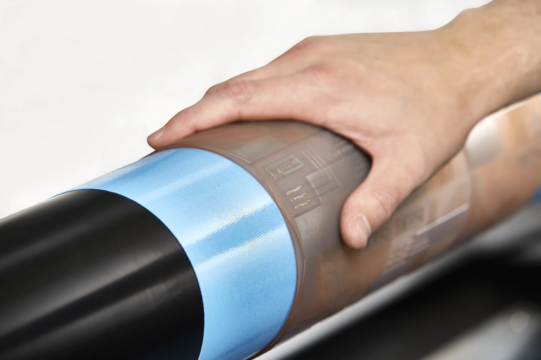 A hand touches a cylindrical roller wrapped with a partially transparent tesa tape, featuring blue and brown sections. The background is blurred and neutral in color. (This text has been generated by AI)