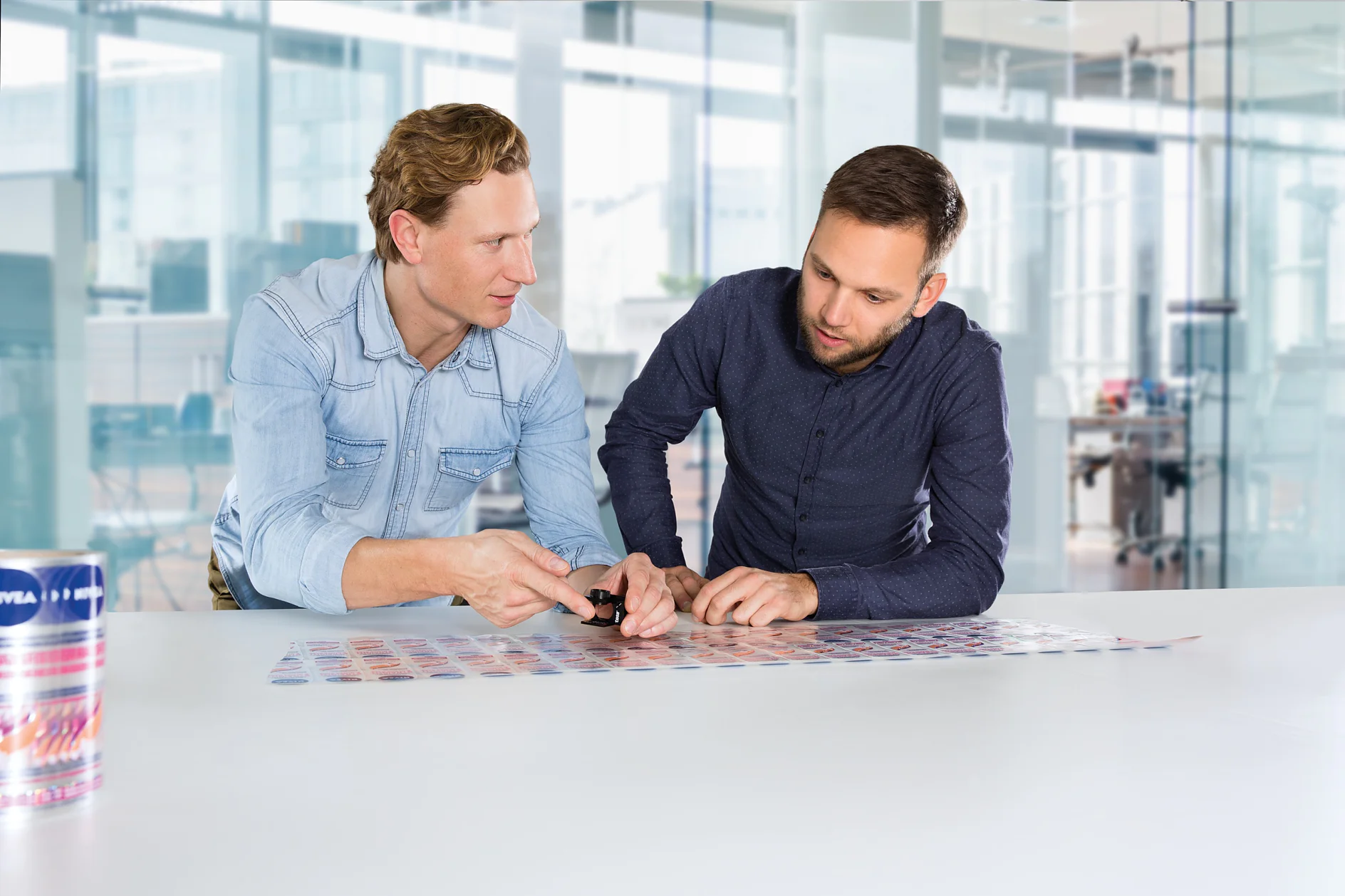 Two men are collaborating at a table in a modern office setting. One man is pointing at a sheet of images on the table, while the other is observing it attentively. A can of soda is visible on the table in the foreground, along with tesa tape nearby. (This text has been generated by AI)