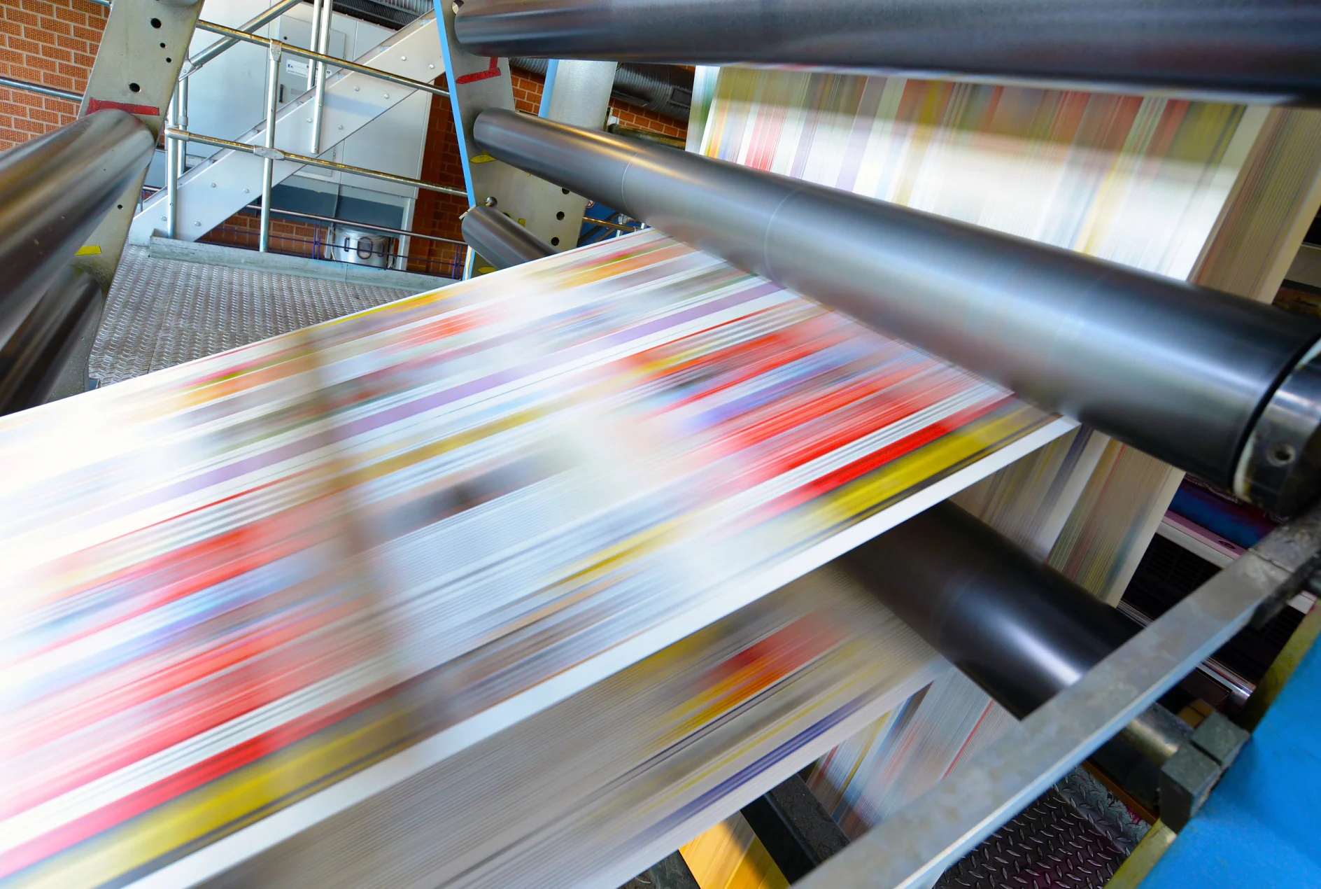 Close-up of a printing press in action, with colorful sheets of paper rapidly moving through rollers. The equipment appears to be part of an industrial printing process, evident from the large scale and speed of operation. (This text has been generated by AI)
