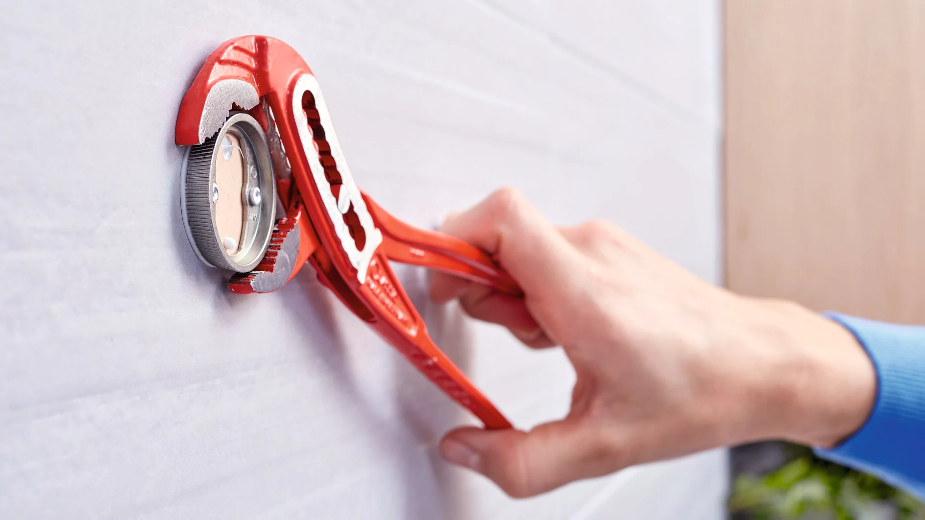 A hand is using a red adjustable wrench to tighten a silver pipe fitting wrapped with tesa tape. The fitting is mounted on a white tiled wall. The close-up shot focuses on the tool and fitting, with a blurred background. (This text has been generated by AI)