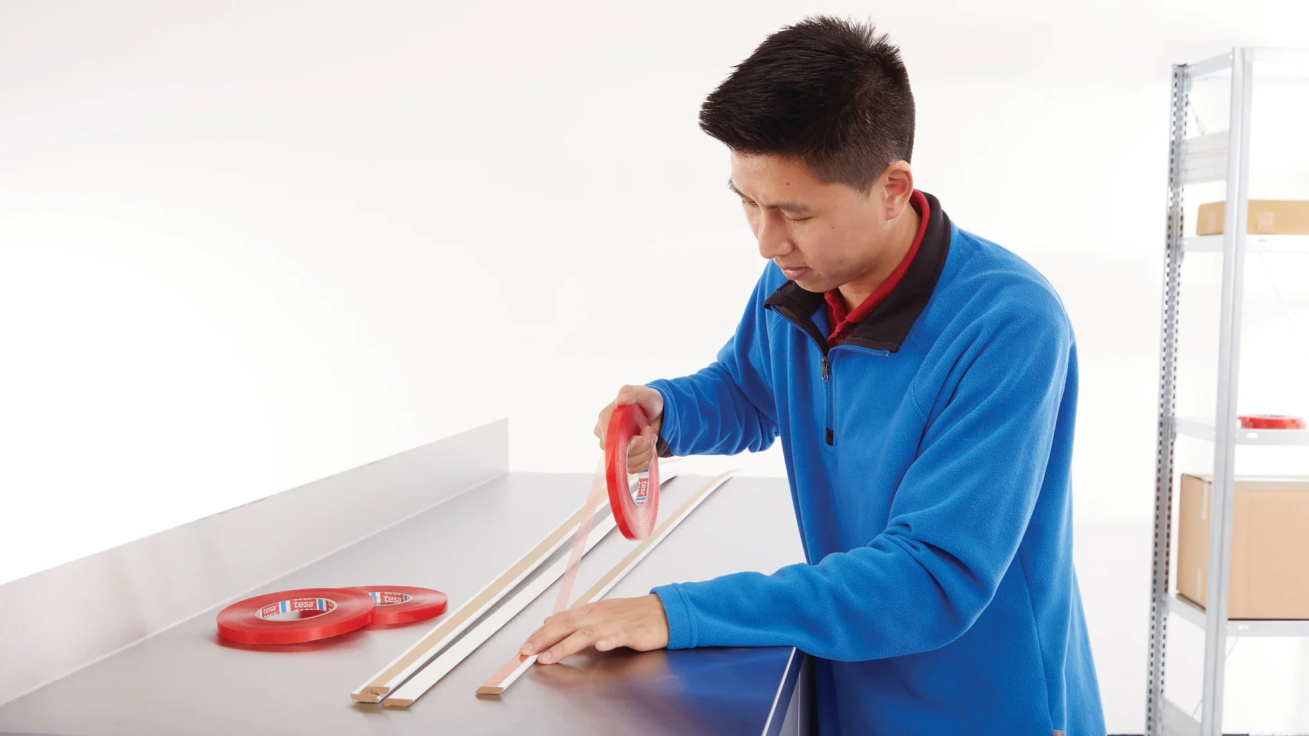 Una persona con una chaqueta azul sostiene un rollo de cinta tesa roja mientras ensambla o mide piezas de madera sobre una superficie metálica. Cerca se ven dos rollos más de cinta tesa. Al fondo, estanterías con cajas. (This text has been generated by AI)