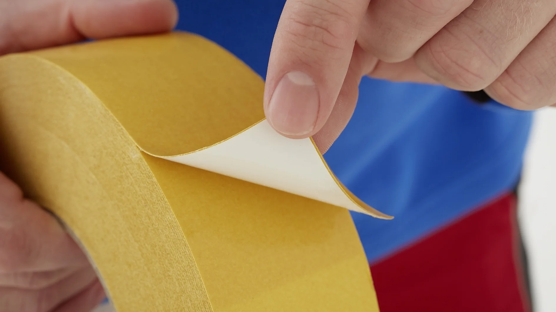 Primer plano de unas manos que despegan una sección de cinta adhesiva de doble cara tesa de un rollo amarillo. Una mano sostiene el rollo mientras que la otra levanta suavemente el soporte protector, dejando al descubierto la superficie adhesiva que se encuentra debajo. (This text has been generated by AI)