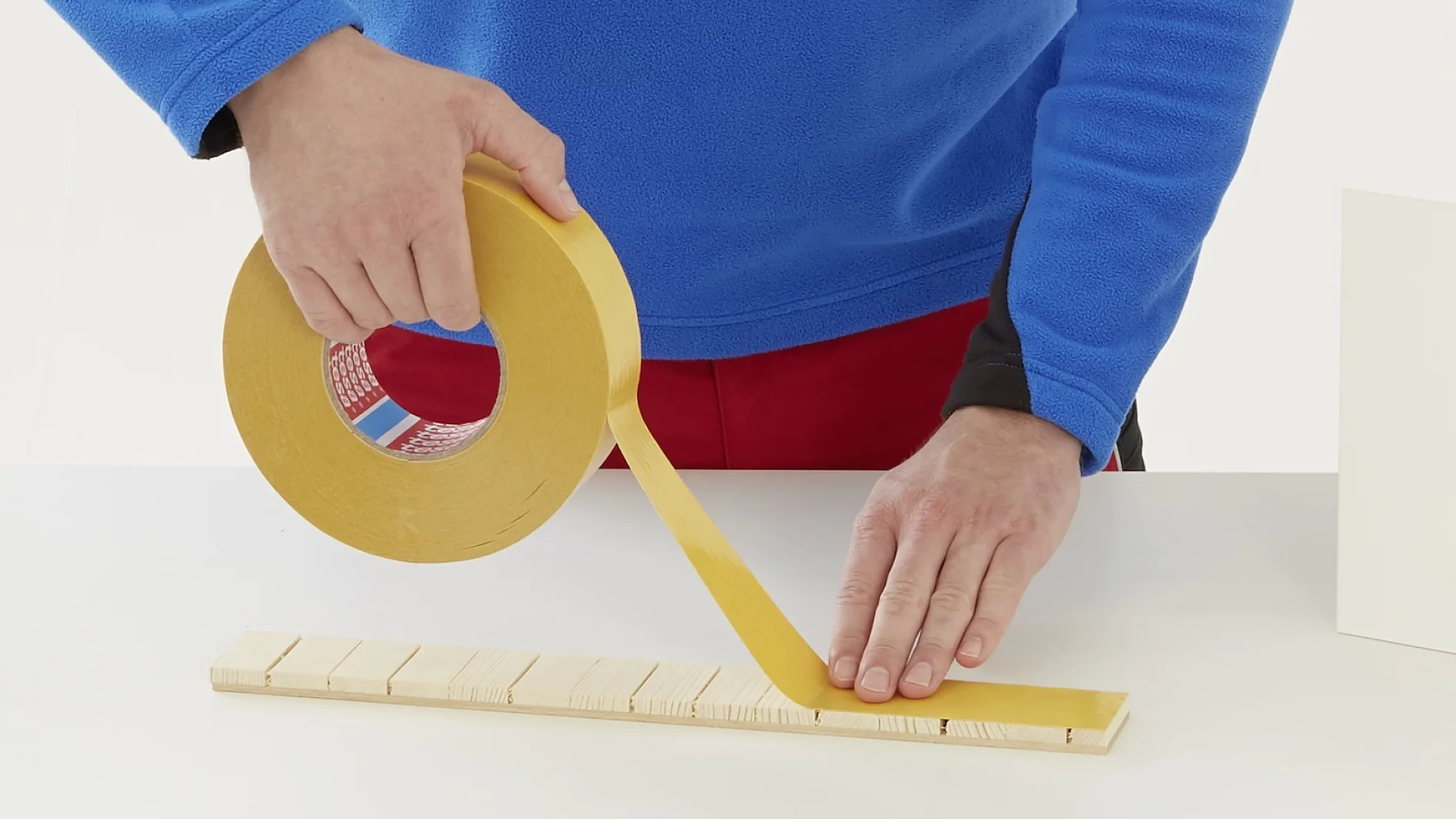Una persona que lleva una chaqueta azul y un pantalón rojo está usando una cinta tesa amarilla ancha para adherirla a una hilera de pequeñas piezas de madera sobre una superficie blanca. Su mano izquierda sostiene la cinta tesa mientras que con la derecha la extiende sobre la madera. (This text has been generated by AI)