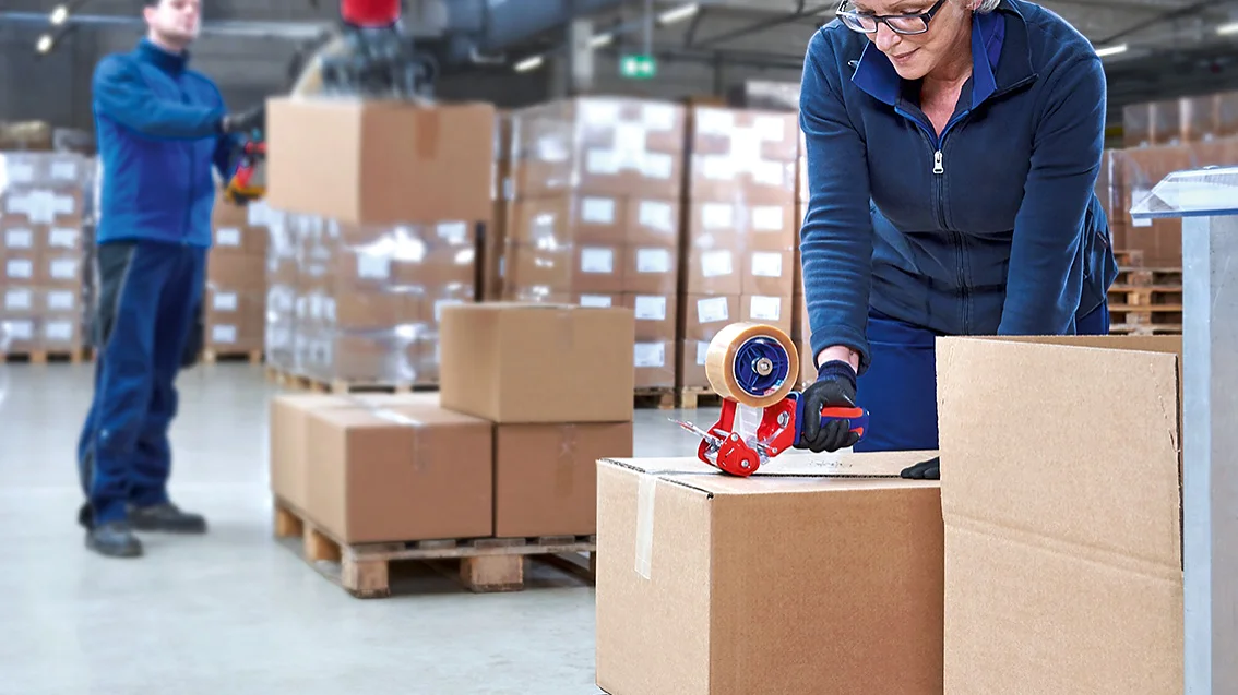 Una mujer de pelo corto y rubio que lleva una chaqueta azul está sellando una caja de cartón con cinta adhesiva Tesa en un banco de trabajo. Un hombre en el fondo, también con chaqueta azul, está manipulando una caja con maquinaria en un almacén. (This text has been generated by AI)