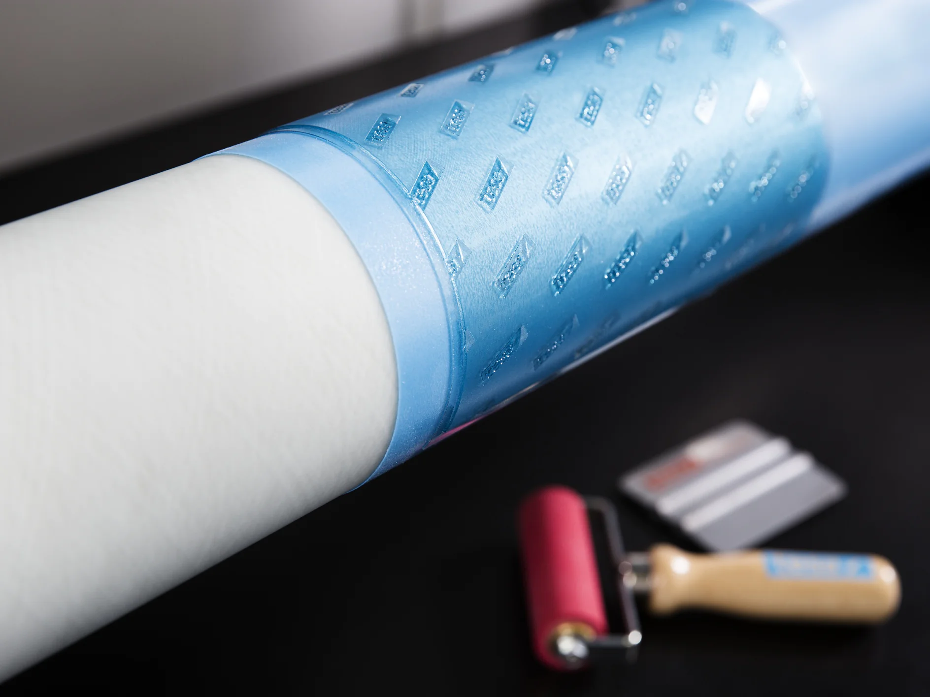 fp-and-p-flexographic-printing-plate-mounting-097 Un rollo de material blanco parcialmente envuelto con plástico texturizado azul. Un rodillo con mango rojo y un pequeño raspador se colocan sobre una superficie negra cercana, junto con cinta tesa. (This text has been generated by AI)
