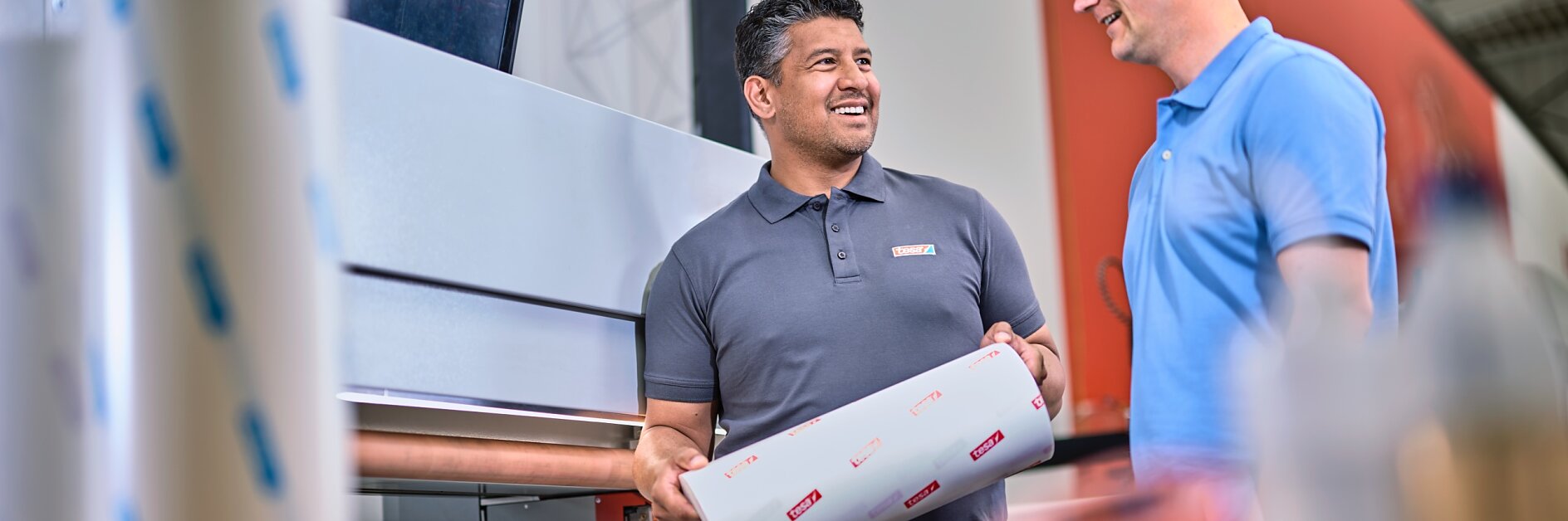 Two men are standing in a printing workshop. One man holds a roll of printed material secured with tesa tape, while the other looks on. A large printer and computer screen are in the background. Both men are dressed in casual work attire.