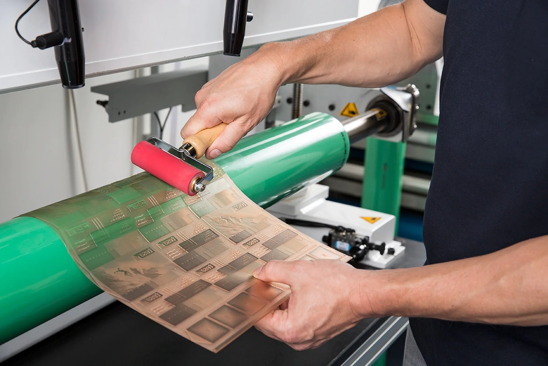 FP&P_Flexographic-Printing_Twinlock-006-Screen Una persona utiliza un rodillo manual sobre una placa de impresión flexográfica. La placa está envuelta alrededor de un gran cilindro verde con cinta tesa. El dispositivo parece ser parte de una máquina de impresión. (This text has been generated by AI)
