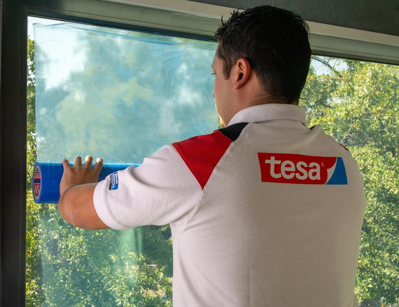 Una persona con una camiseta de la marca Tesa aplica una película de cinta adhesiva azul Tesa a una ventana grande. La vista exterior muestra árboles verdes y un cielo despejado. (This text has been generated by AI)