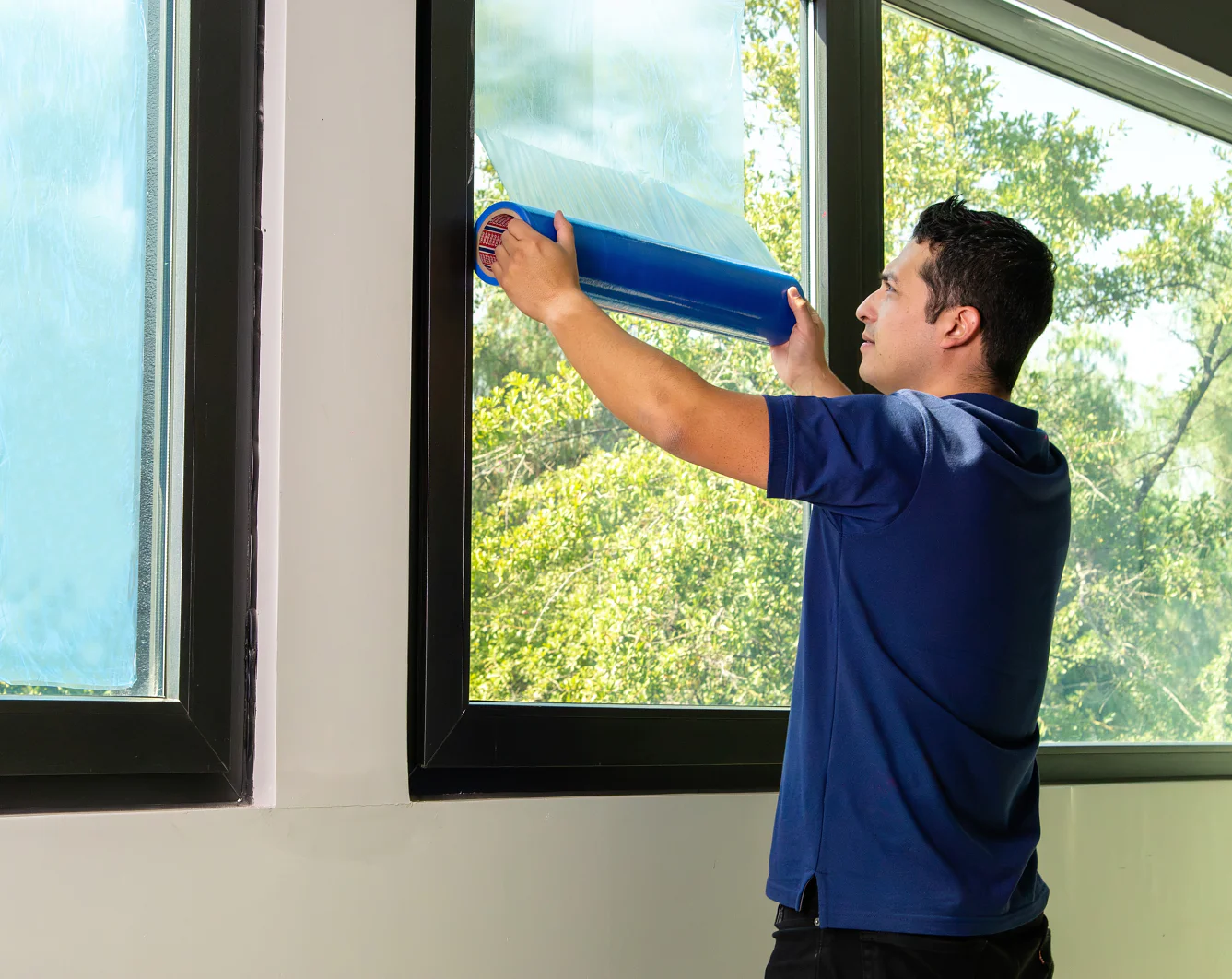 Una persona con una camisa azul aplica una película azul sobre una ventana grande. La escena se desarrolla en un interior con árboles visibles en el exterior. La persona está concentrada en desenrollar la cinta tesa, de pie cerca de una pared. (This text has been generated by AI)
