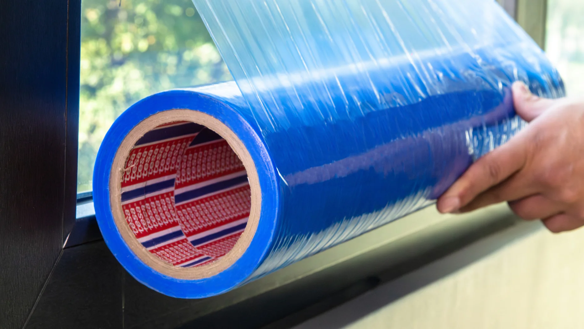 Una persona desenrolla un rollo grande de cinta adhesiva azul tesa cerca de una ventana. La cinta tesa presenta un patrón rojo y blanco en el interior del rollo. La ventana parece parcialmente cubierta y se ven árboles afuera. (This text has been generated by AI)