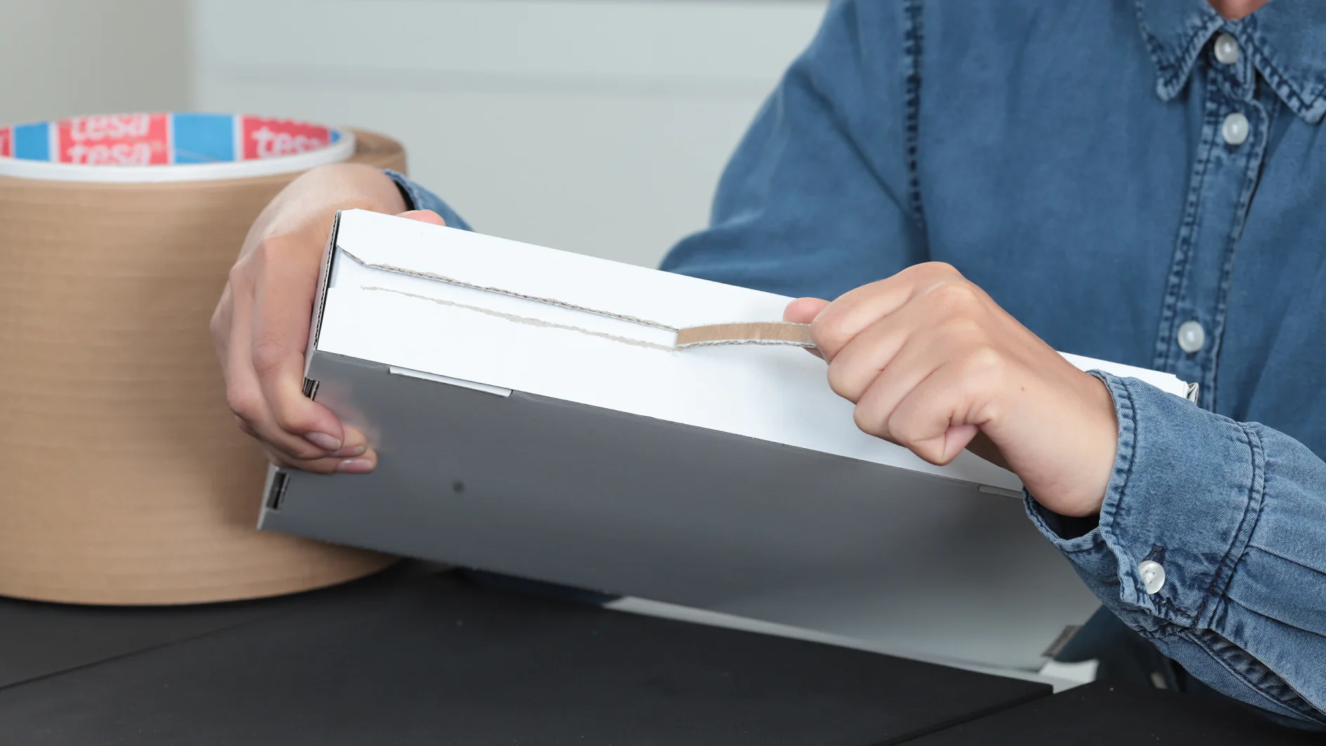 A person in a blue shirt is opening a cardboard box by pulling a tab. A roll of tesa tape is visible in the background on a black table. (This text has been generated by AI)