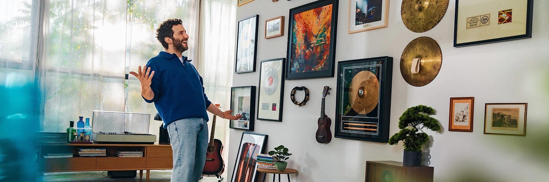 Un hombre con sudadera azul y vaqueros sonríe con los brazos abiertos en una habitación luminosa, frente a una pared decorada con cuadros, instrumentos musicales, platillos y plantas, todo ello fijado con cinta adhesiva. Al fondo se ven un tocadiscos y una guitarra. (This text has been generated by AI)