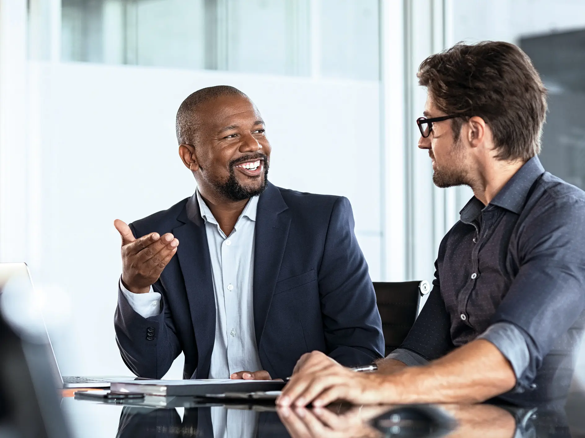 Two men in business attire engaged in discussion at an office desk with laptop and documents, indoor professional setting. (This text has been generated by AI)