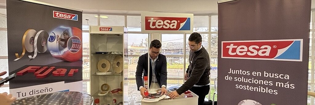 Dos hombres en un stand de tesa y Microgeo, rodeados de pancartas, productos de cinta adhesiva de tesa y una estantería de exposición en un luminoso espacio interior.