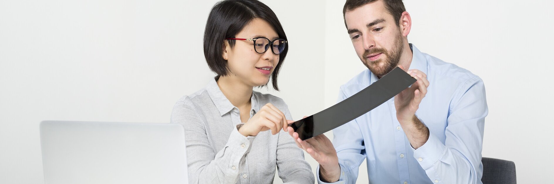 Dos personas en un escritorio blanco con un portátil y documentos examinan una cinta tesa negra flexible, ambas enfocadas mientras una apunta y otra la sujeta. (This text has been generated by AI)