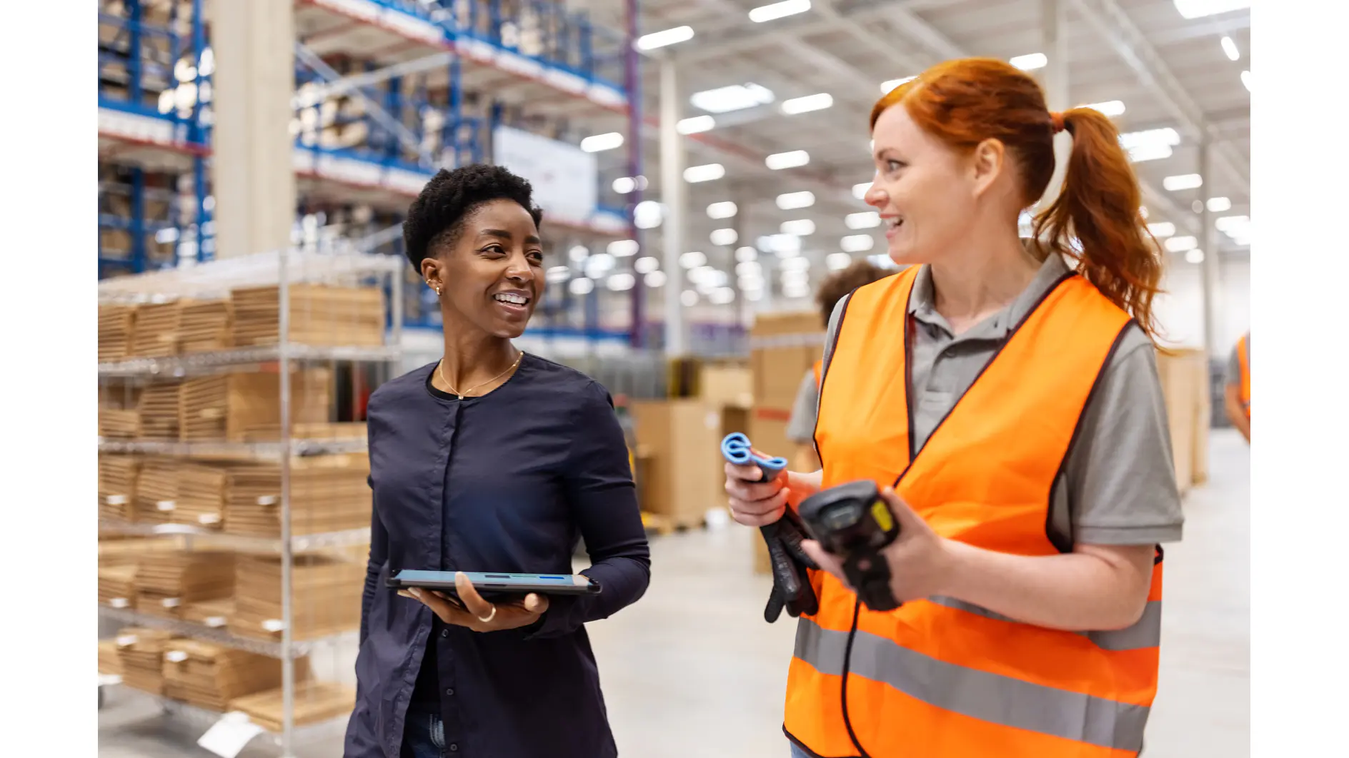 Two women in a warehouse talk while walking; one holds a tablet and wears business attire, the other wears a high-visibility vest and holds a barcode scanner. Shelves with boxes—and prominently displayed rolls of tesa tape—are visible in the background. (This text has been generated by AI)