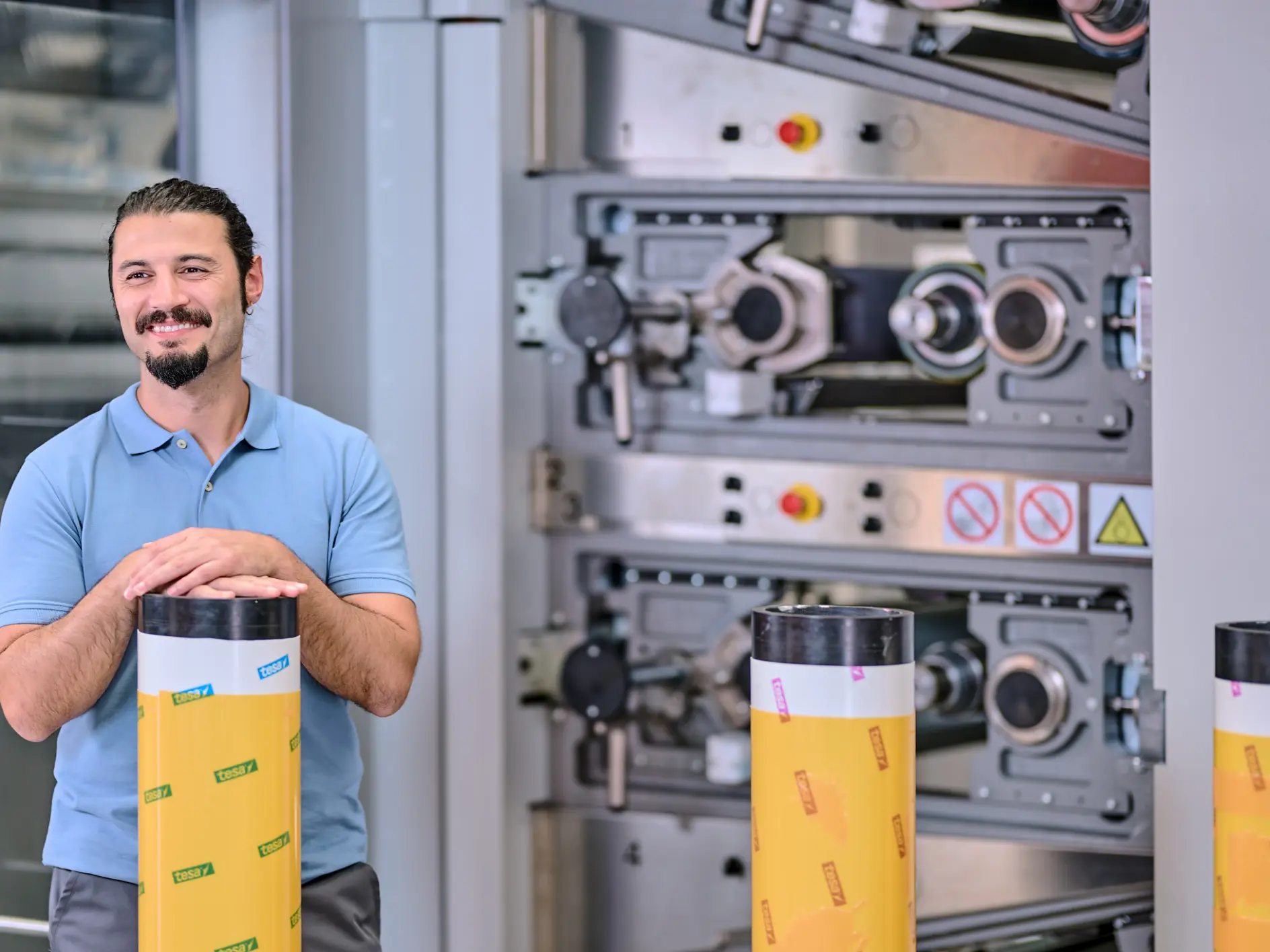 Un hombre con barba y cola de caballo, que lleva puesta una camisa tipo polo azul, está de pie sonriendo con las manos sobre un gran objeto cilíndrico envuelto en cinta tesa amarilla. En el fondo se puede ver maquinaria industrial. (This text has been generated by AI)