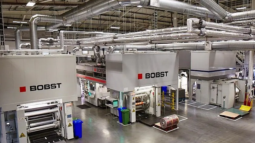 Una gran instalación industrial con múltiples máquinas Bobst para impresión o embalaje. Las máquinas son blancas con logotipos rojos y negros. La sala tiene un techo alto con ductos expuestos e iluminación superior. Se pueden ver varios materiales, incluyendo cinta tesa, y contenedores. (This text has been generated by AI)