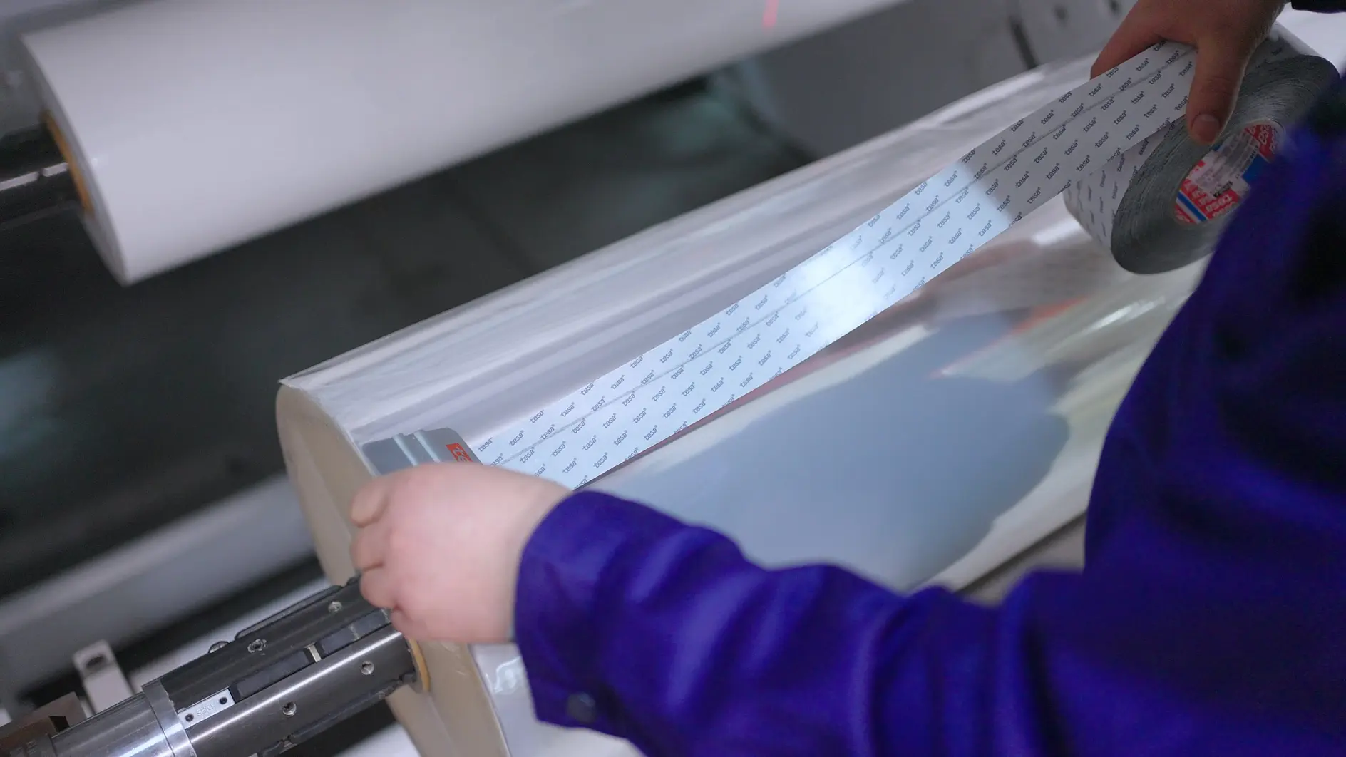 A person in a blue shirt is holding a roll of clear tesa tape with printed labels. The roll is mounted on a machine, likely for processing or packaging. The background is blurred, focusing on the roll and hands.