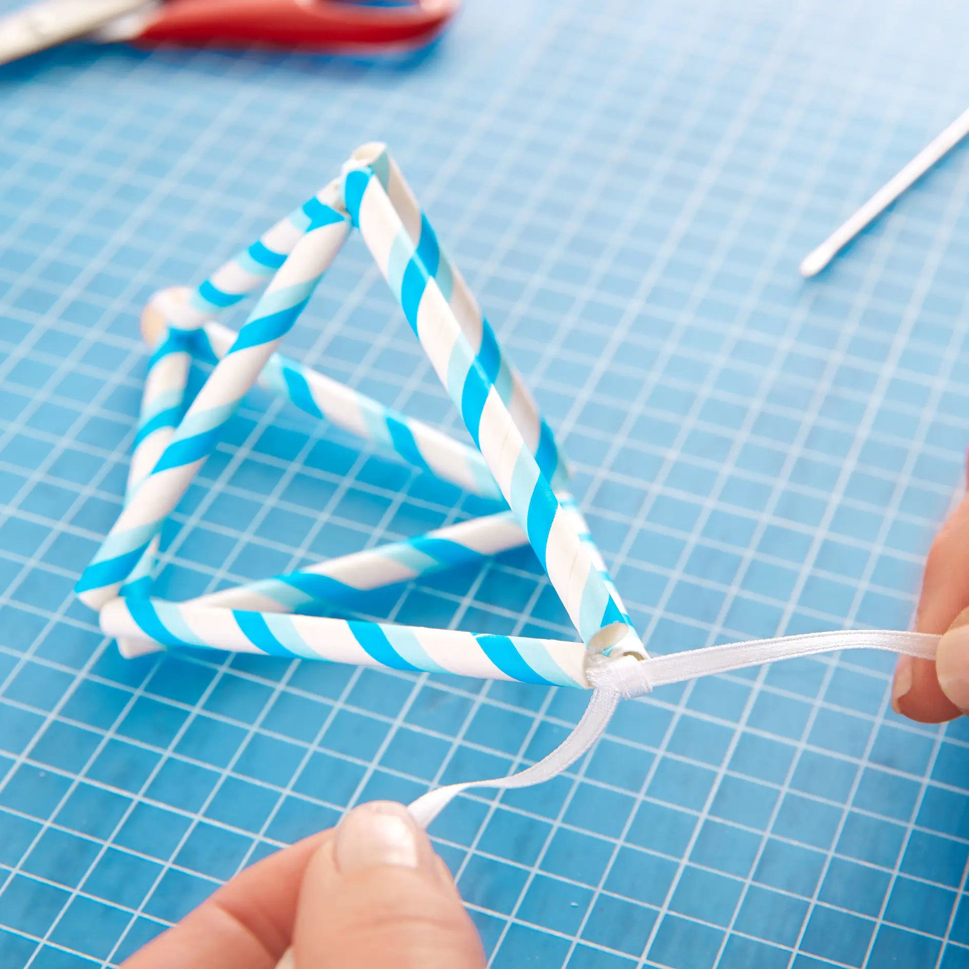 Close-up of hands tying a white ribbon to a geometric structure made of blue and white striped paper straws, forming a triangle. The structure rests on a blue grid-patterned surface, with a pair of scissors in the background. (This text has been generated by AI)