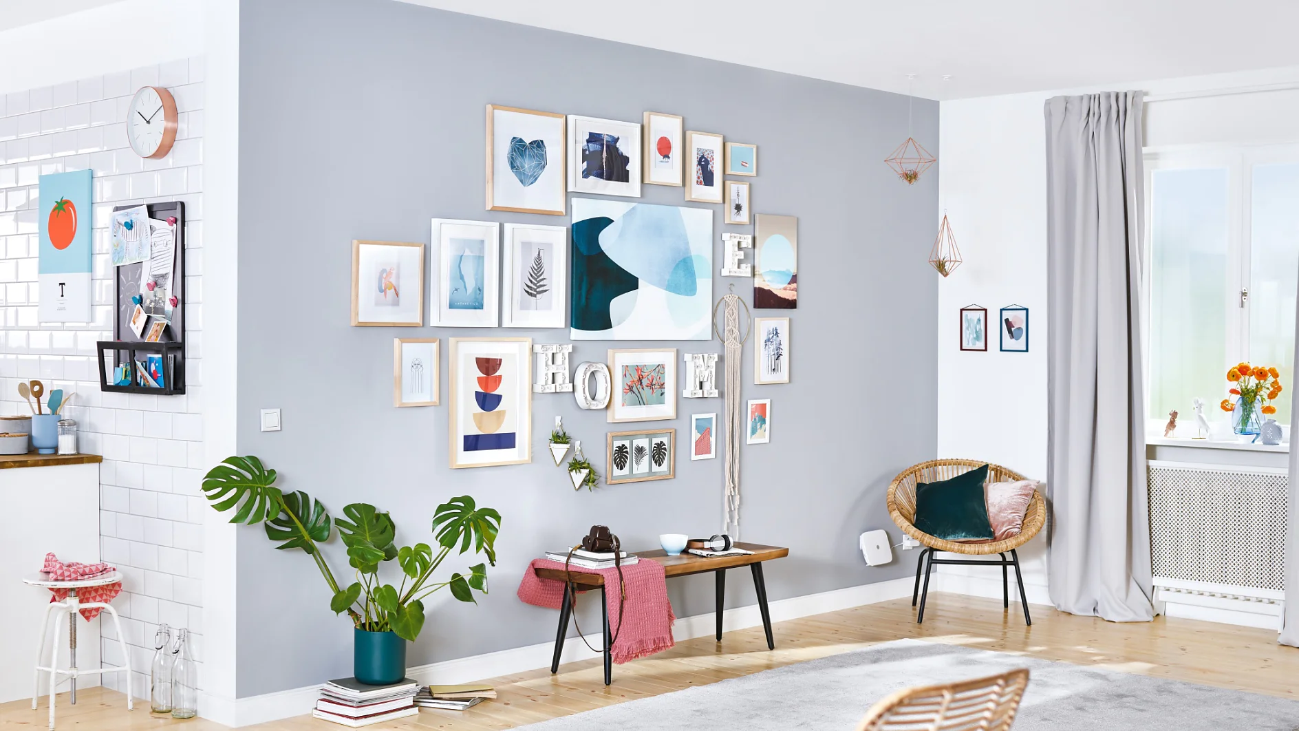 A modern living room with a light gray accent wall featuring a collage of framed art, all expertly mounted with tesa tape. The room includes a wooden bench, two chairs, a plant, and light gray curtains. The adjacent kitchen area boasts white tiles and wooden countertops. (This text has been generated by AI)