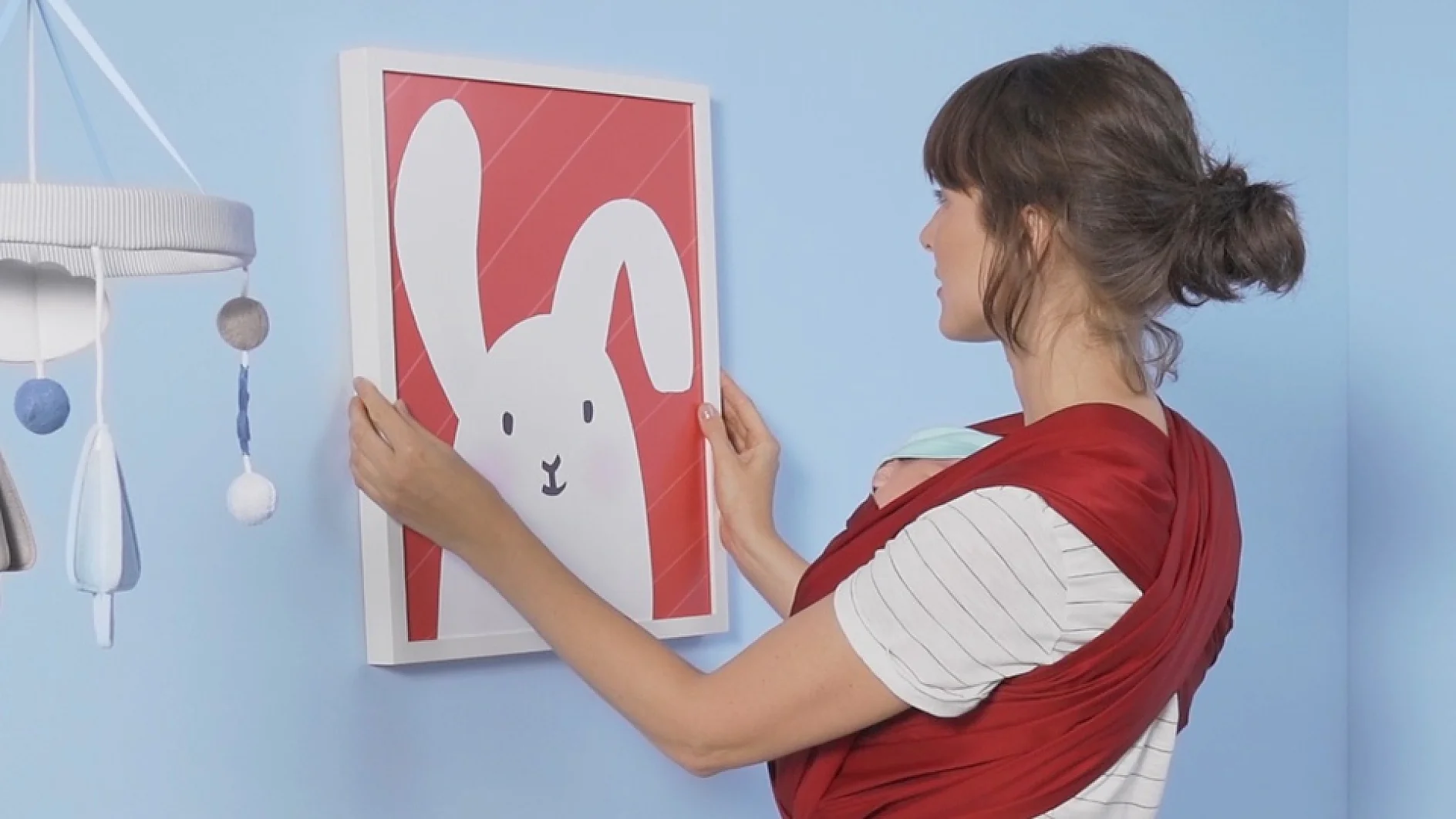 A woman in a striped shirt stands against a light blue wall, adjusting a framed picture of a white cartoon bunny on a red background. She is wearing a red baby carrier with a sleeping baby nestled inside. A mobile with soft shapes hangs nearby, secured using tesa tape. (This text has been generated by AI)