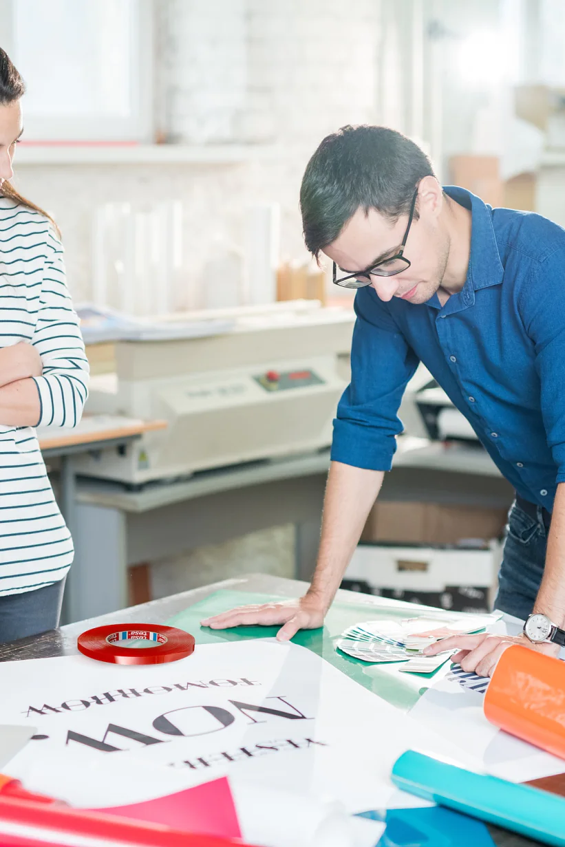 Two people in a print shop examine a large poster on a table. The man is wearing glasses and a blue shirt, while the woman is in a striped shirt with her arms crossed. The table is cluttered with rolled-up paper, tesa tape, and a laptop. (This text has been generated by AI)
